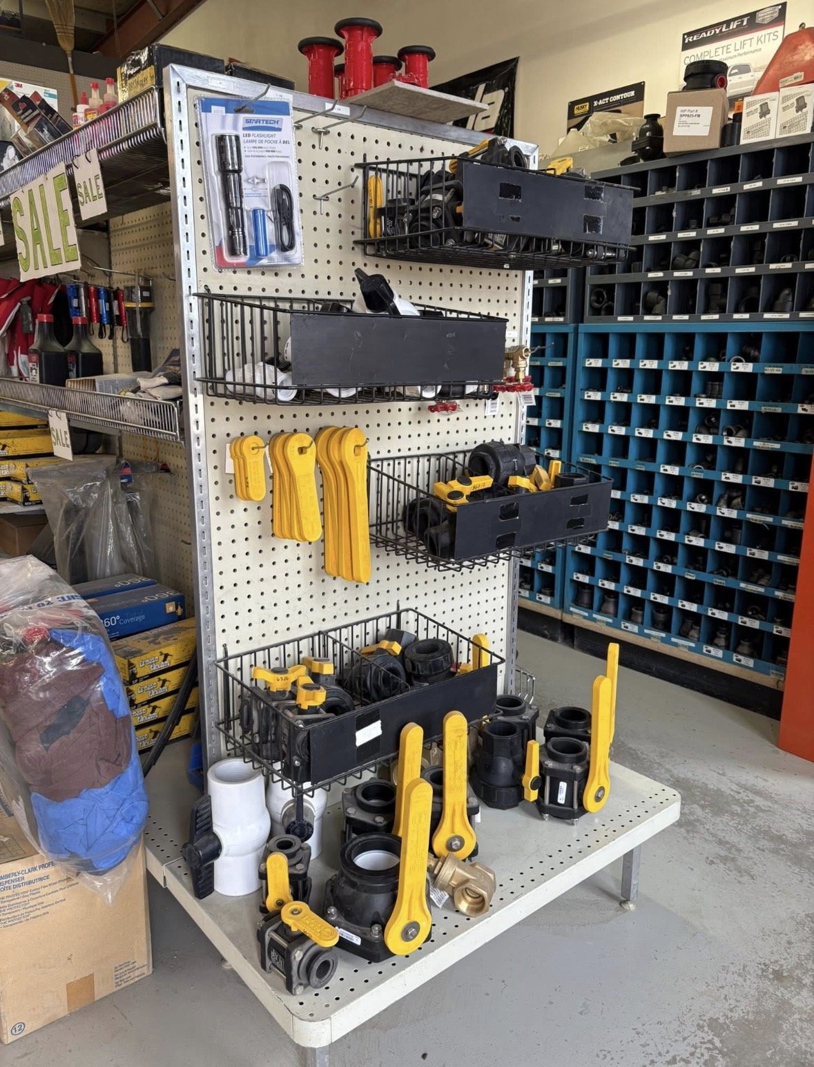 Display of various plumbing and hardware fittings, including pipe connectors, valves, and yellow safety locks, on a pegboard and table at a hardware store.