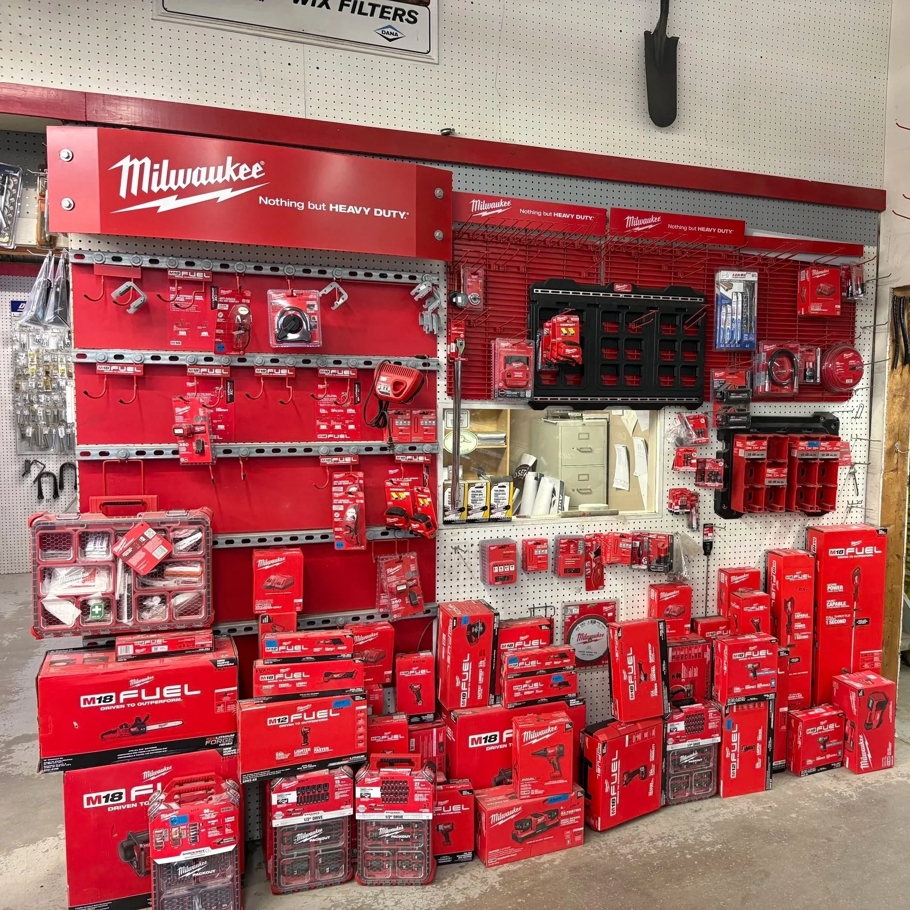 Display of Milwaukee power tools and accessories, including batteries, chargers, and tool kits, arranged on a pegboard at a hardware store.