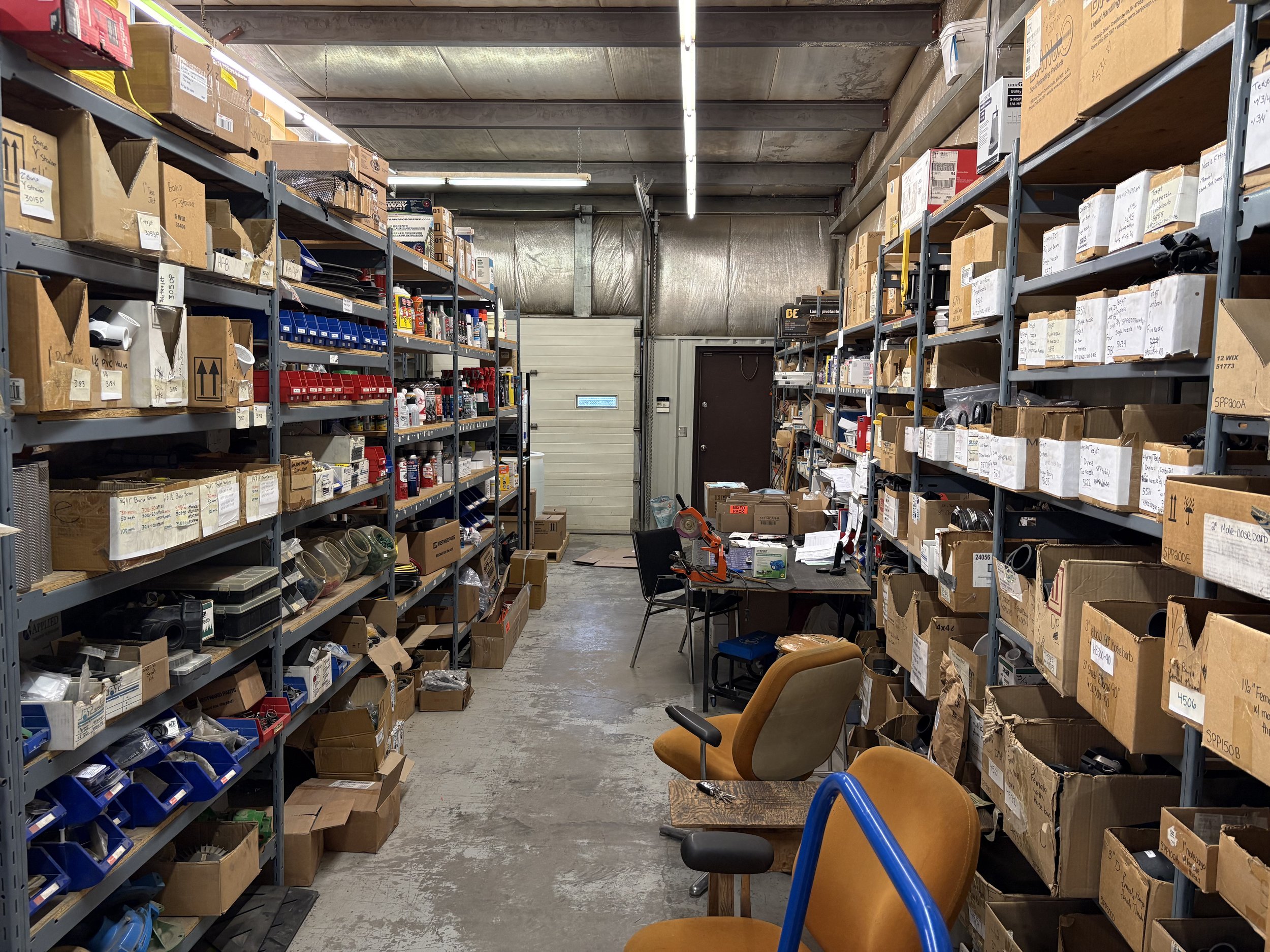 A cluttered storage room with metal shelves filled with labeled cardboard boxes and plastic bins, a small work table with tools, and wooden chairs. The room has an industrial ceiling with fluorescent lights and a metal garage door at the back.