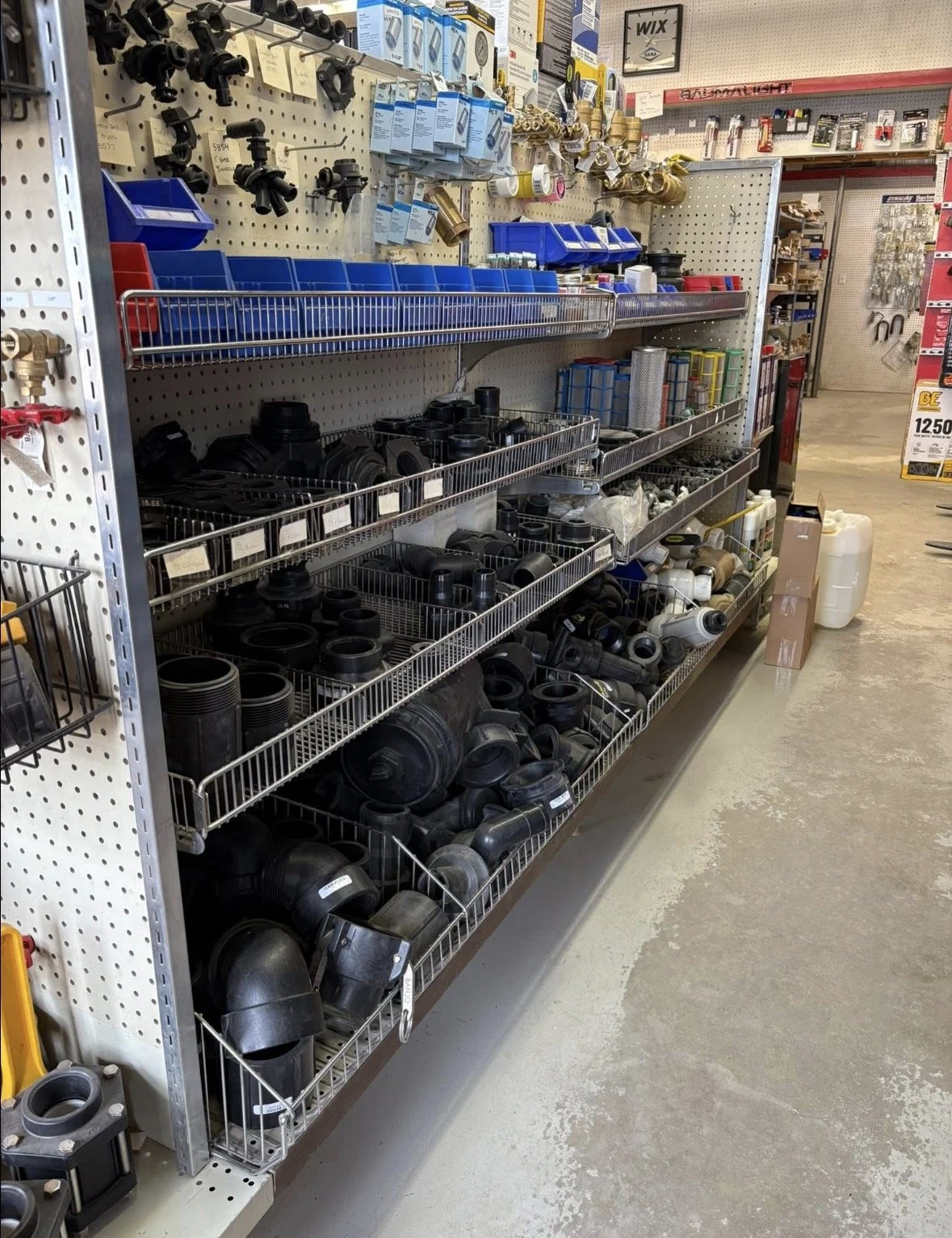 Store aisle with shelves stocked with plumbing parts, fittings, and hardware in bins and on pegboard.