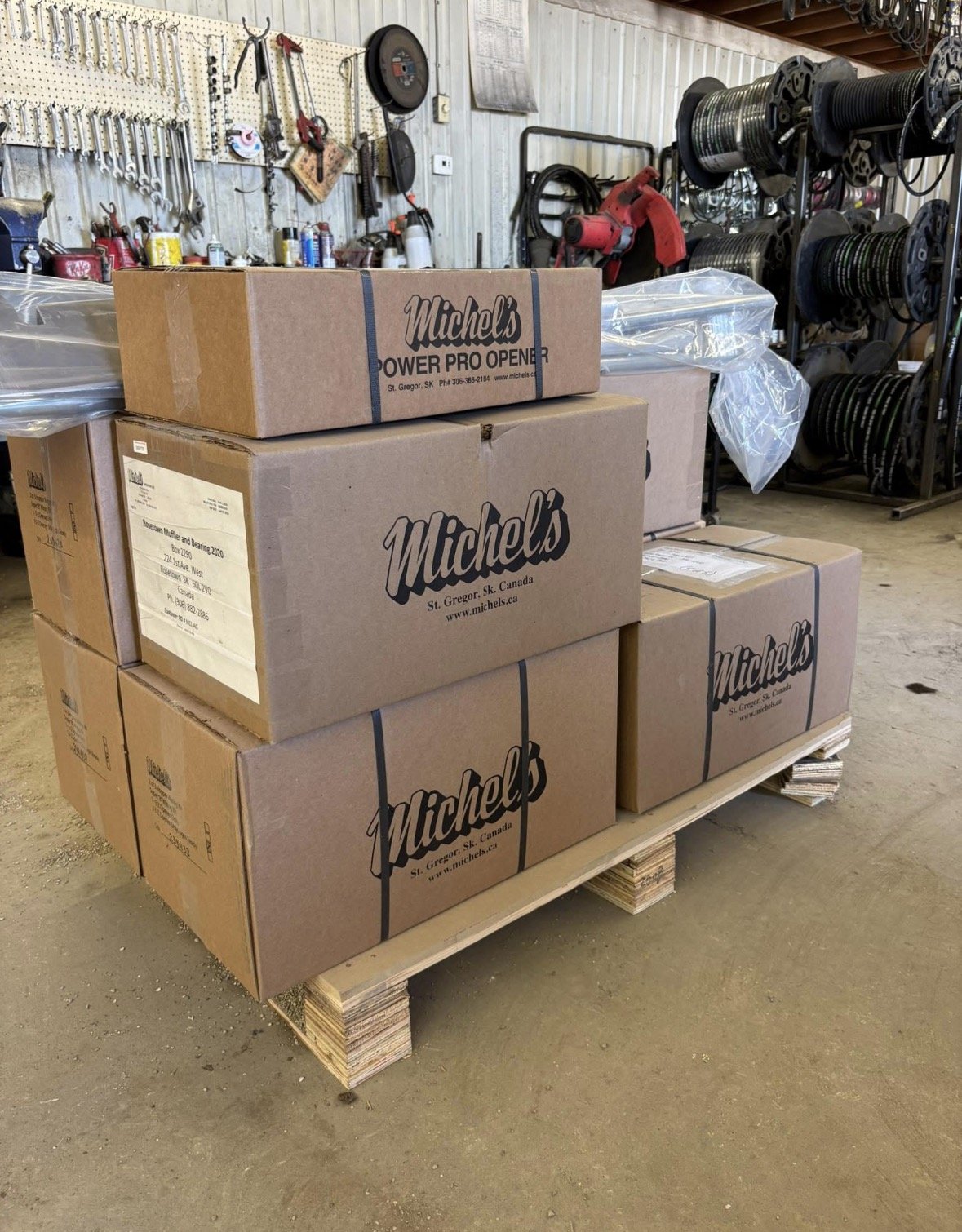 St.urdy boxes labeled 'Michels' stacked on a wooden pallet in a workshop or warehouse setting with various tools and equipment hanging on the wall.