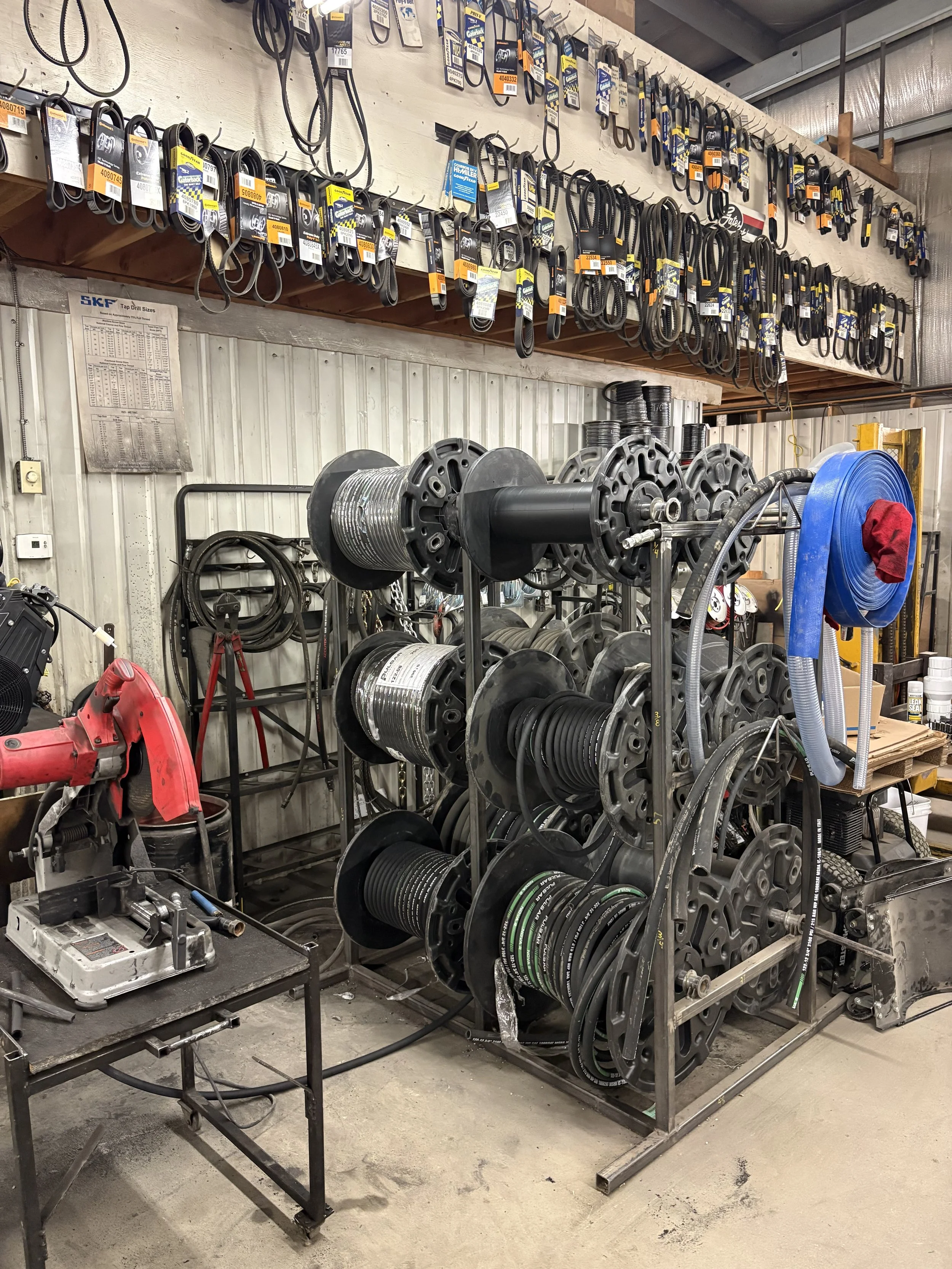Workshop or industrial space with metal shelving holding various belts and hoses, a workbench with tools, and a large spooled hose or cable on a stand.