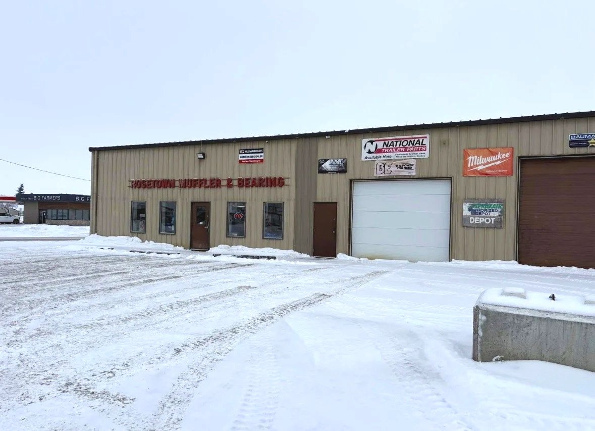 A beige metal building with red sign reading "ROSETOWN MUFFLER & BEARING" on a snowy day. Several signs are posted on the building, including "National Trailer Parts" and "Milwaukee." The ground is covered in snow with visible tire tracks, and there are small snow piles near the building.