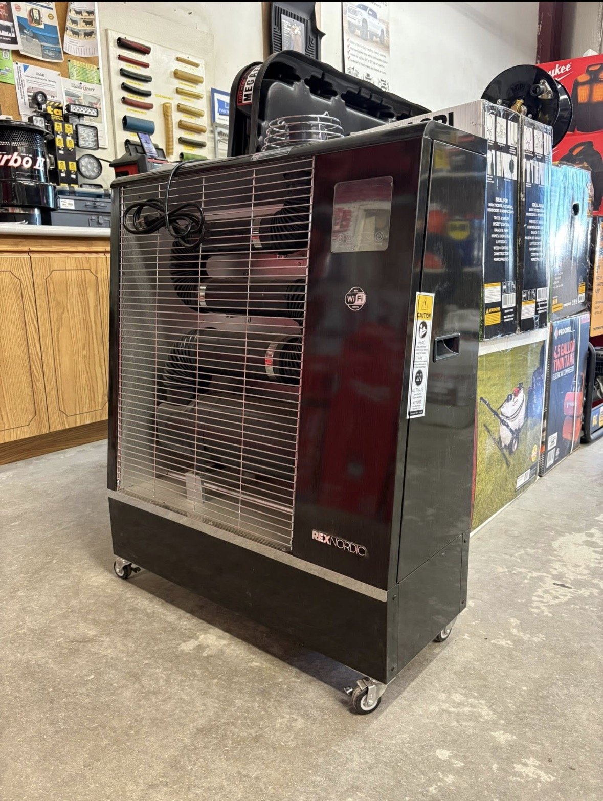 A large black space heater with the brand name Rex Nordic on the bottom front and fractal metal grill covering the front. The heater has four wheels at the bottom for mobility and is located in a hardware store or workshop with various tools and equipment visible in the background.