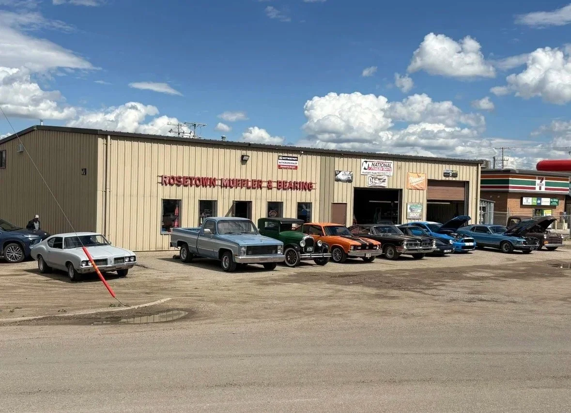 A row of vintage and modern cars parked outside a beige auto repair shop with a sign that reads 'Rosetown Muffler & Bearing' under a blue sky with white clouds.