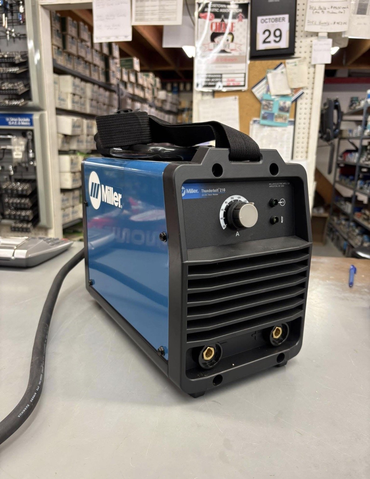 A Miller Thunderbolt 210 welding machine with a black control panel, a dial, and ventilation slats, placed on a table in a hardware store with shelves of supplies and a cork bulletin board in the background.