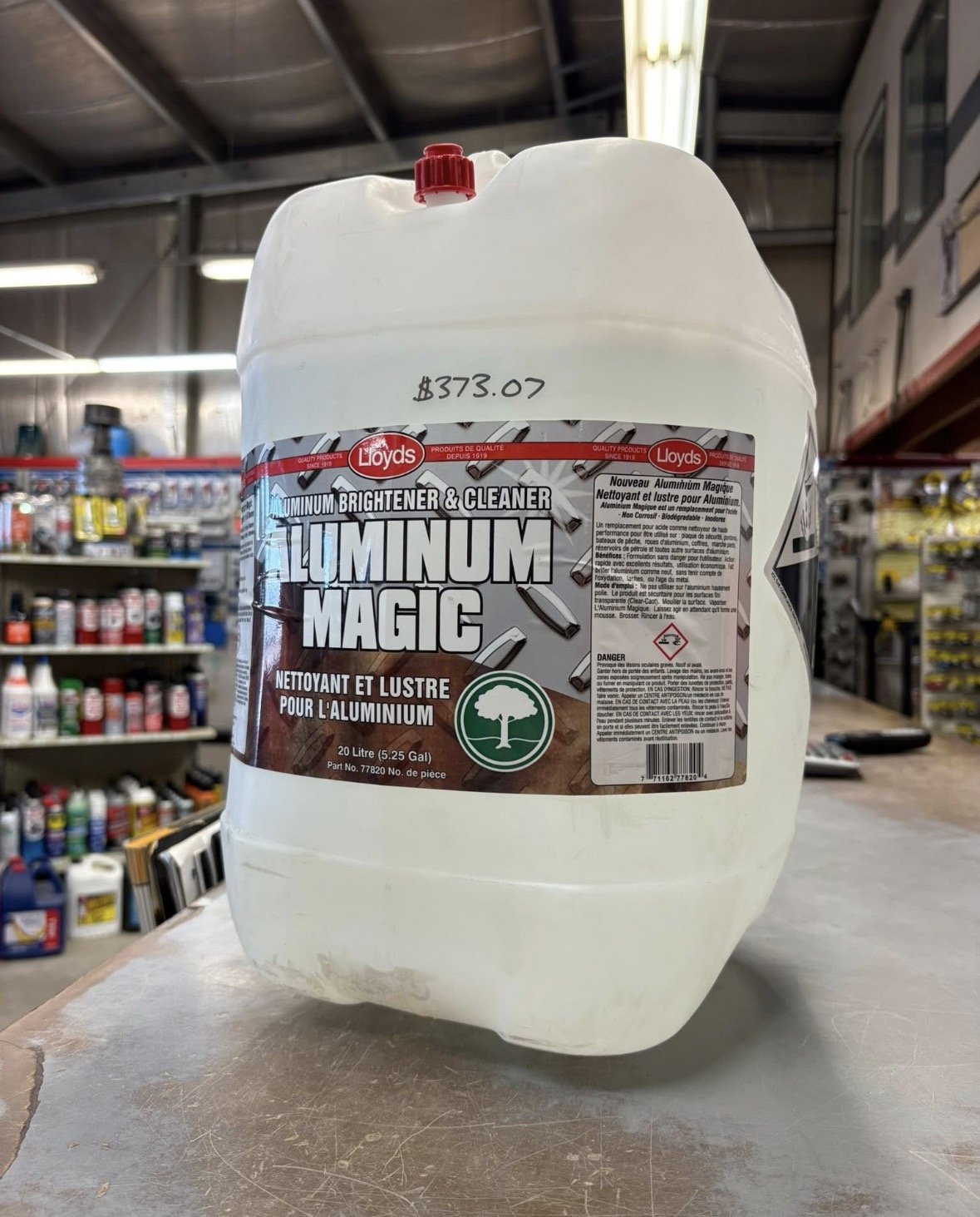 A large white plastic container of Lloyds Aluminum Brightener & Cleaner, labeled as Aluminum Magic, 20 liters, placed on a wooden surface in a hardware store aisle with shelves of paint and other supplies in the background.