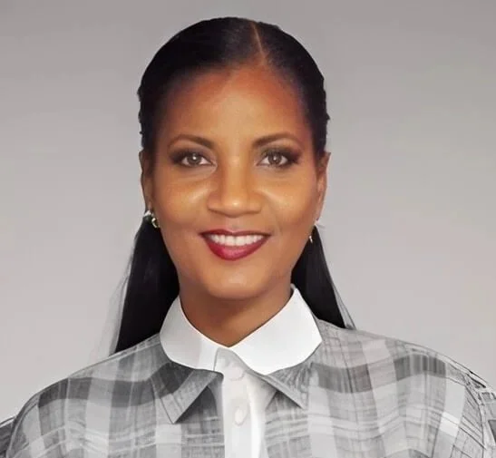 Professional portrait of Sylvia Henry, a woman with long black hair, wearing a white collared shirt and a gray checkered blazer, smiling against a plain gray background.