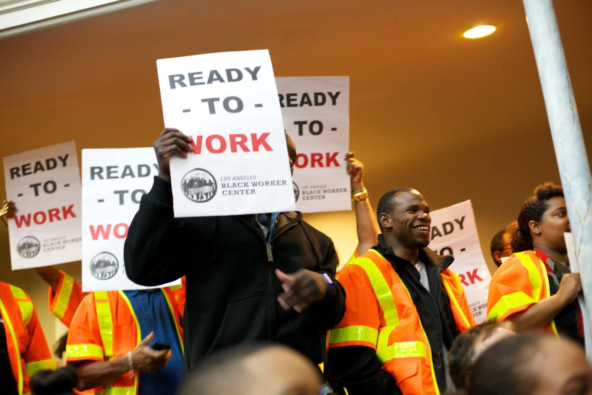 A diverse group of people in safety vests protesting with signs that read "Ready to Work" during a demonstration, with a ceiling and a pole in the background.