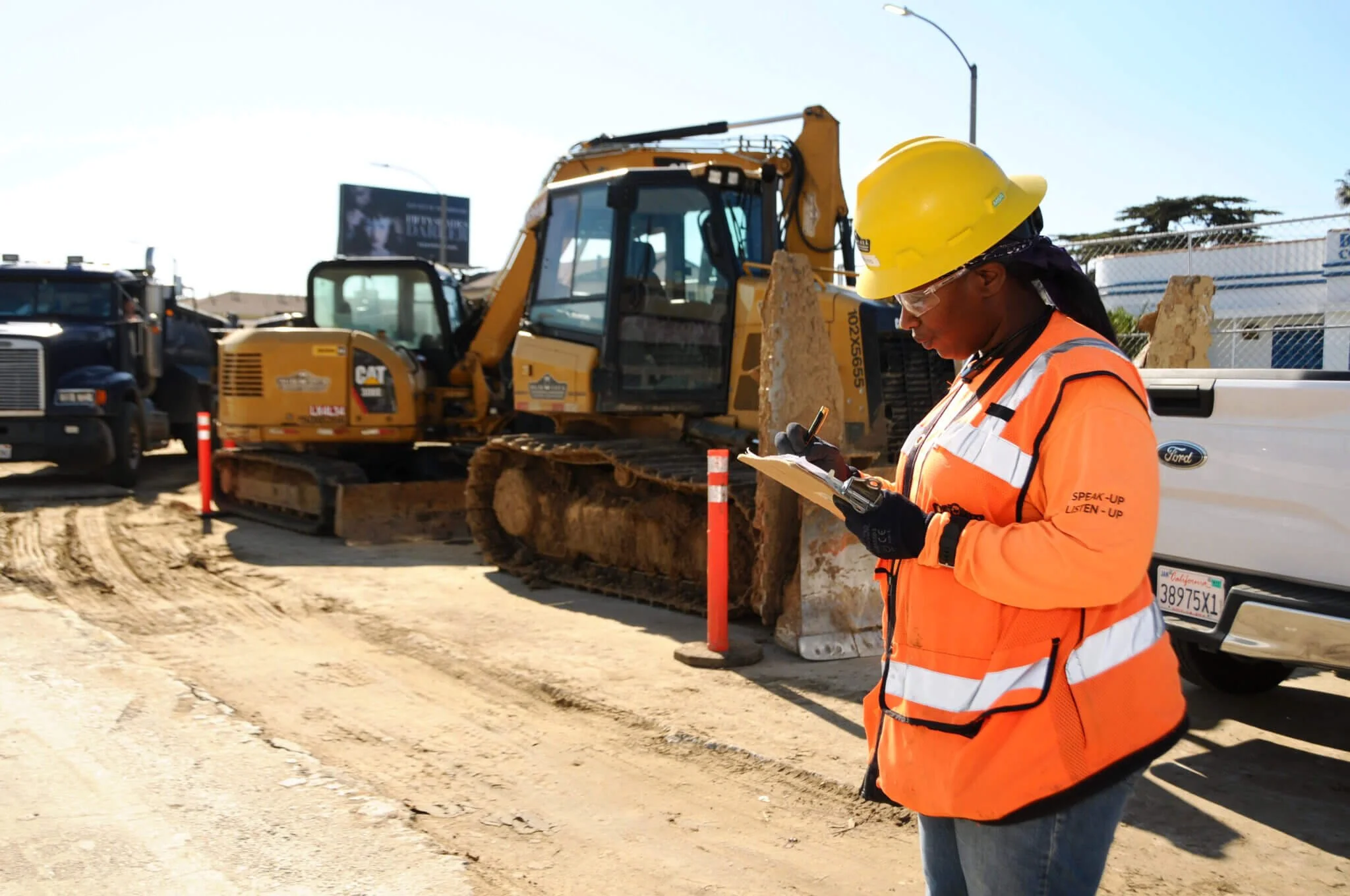Construction worker wearing a yellow safety helmet, safety glasses, and an orange safety vest, writing on a clipboard at a construction site with heavy equipment and vehicles in the background.