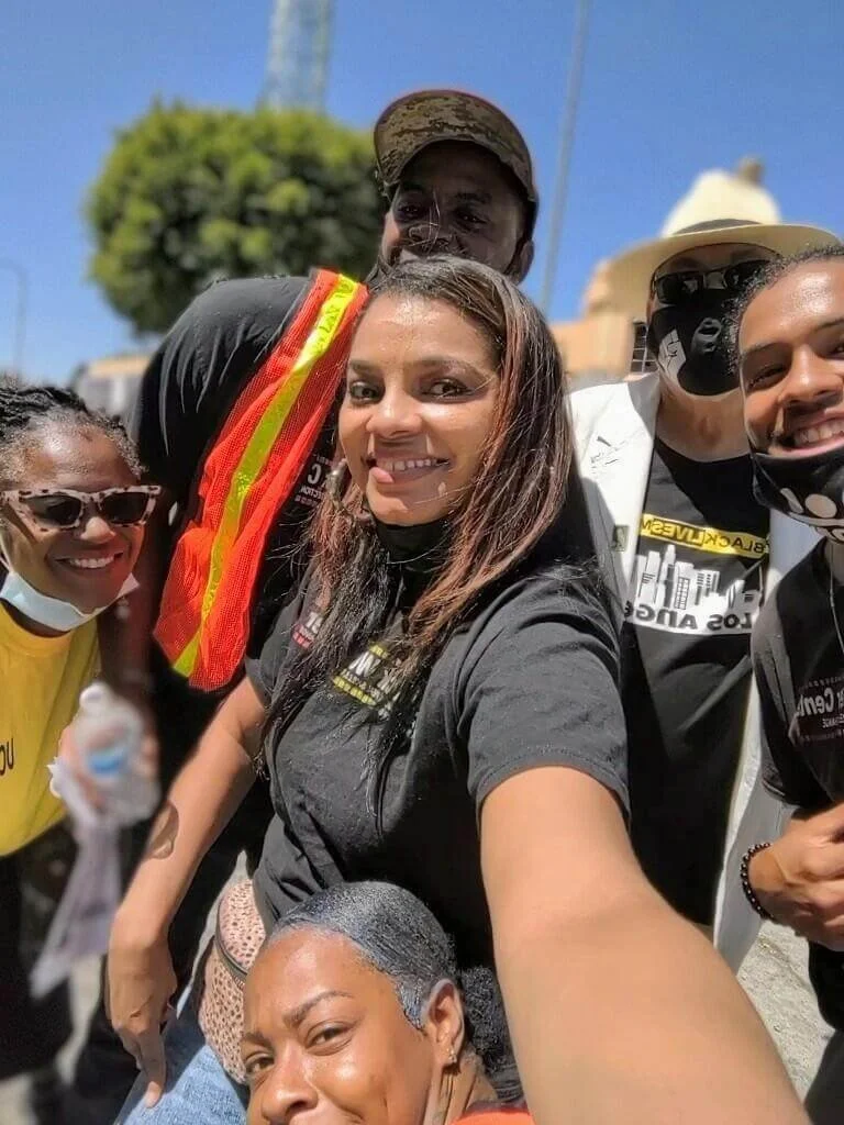 A group of six diverse people smiling and taking a selfie outdoors on a sunny day, with trees and a building in the background.