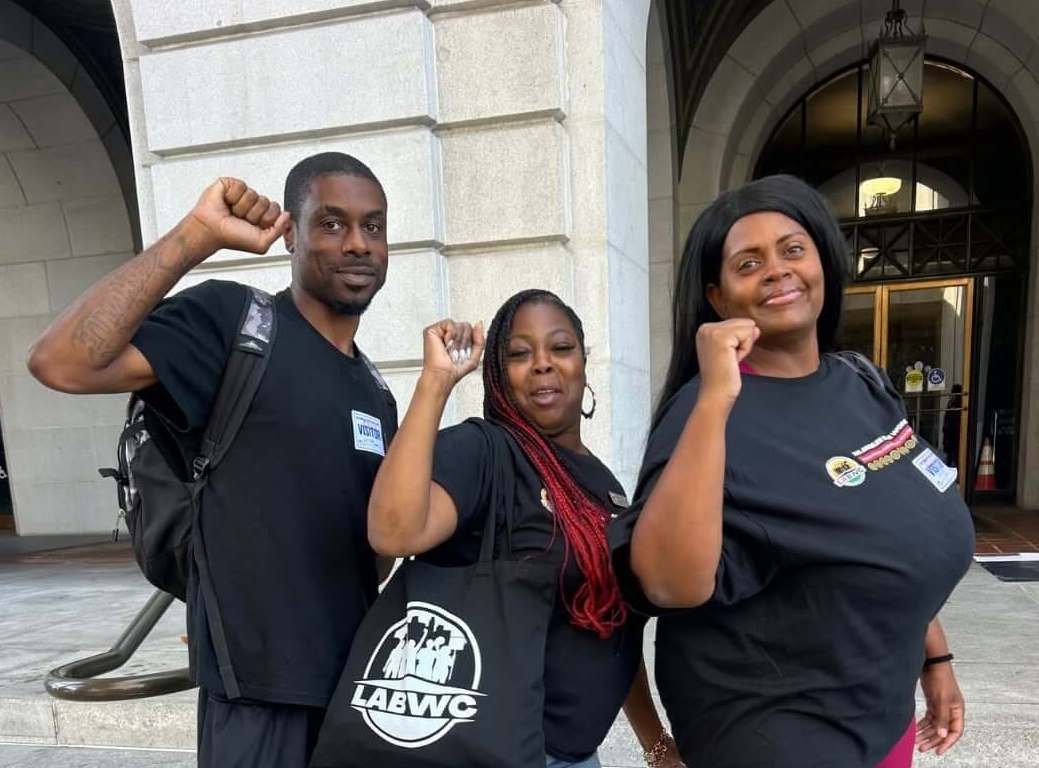 Three people standing outside a building, raising clenched fists. They are dressed in black T-shirts with badges, and have buttons on their shirts. They are smiling and appear to be part of a protest or rally.