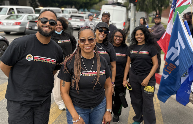 Group of smiling people wearing black T-shirts standing outdoors at a May Day protest, with flags and cars in the background.