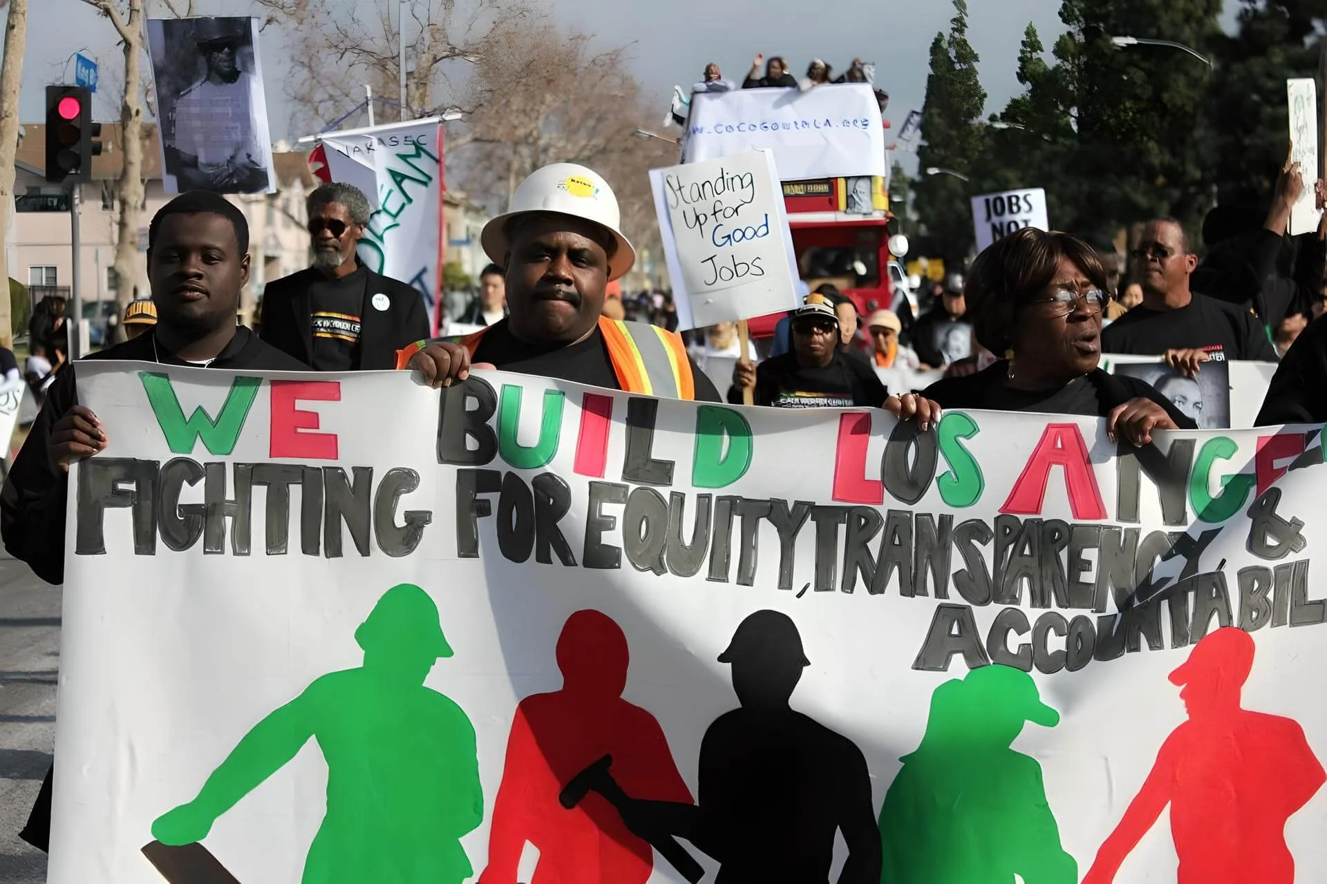 Protesters participating in a march, holding signs and banners advocating for fighting for equity and transparency. Some signs include messages like 'Standing up for good jobs' and 'Jobs now.' The protesters are diverse, and some have silhouettes of people in bright colors on the banner.