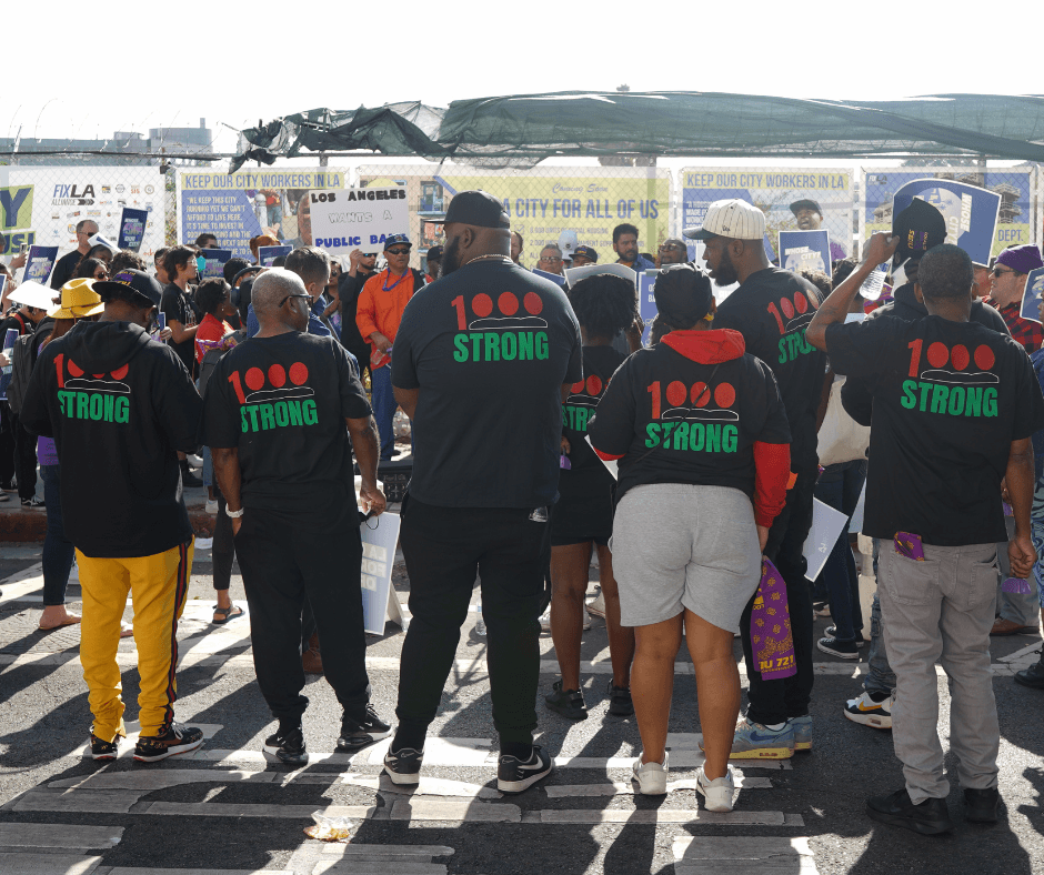 Group of people participating in a rally or protest, wearing matching shirts that say '1000 STRONG' and holding signs supporting city workers in Los Angeles.