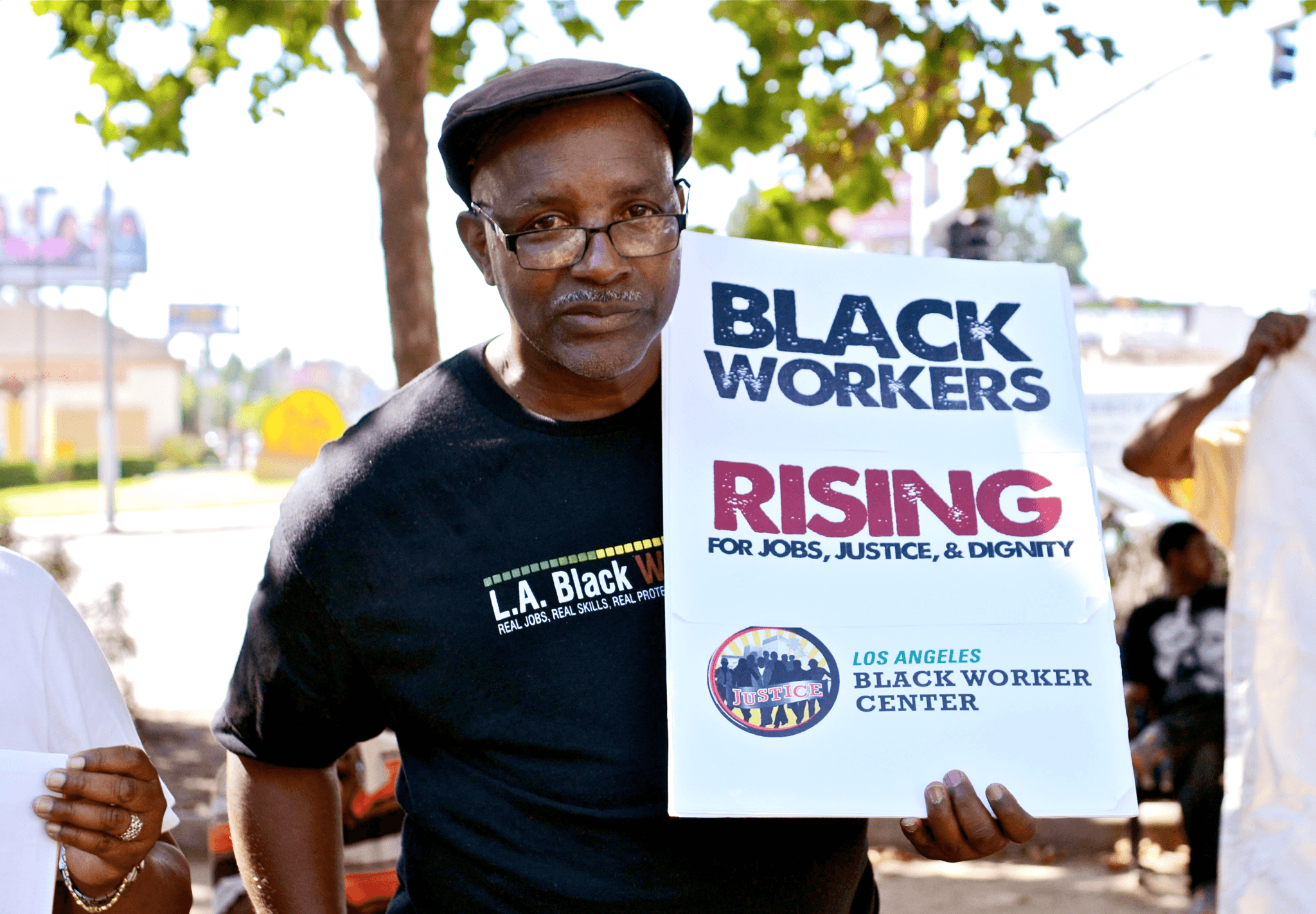 A Black man holding a protest sign that reads, "Black Workers Rising" and "Los Angeles Justice for Black Worker Center" during an outdoor demonstration.