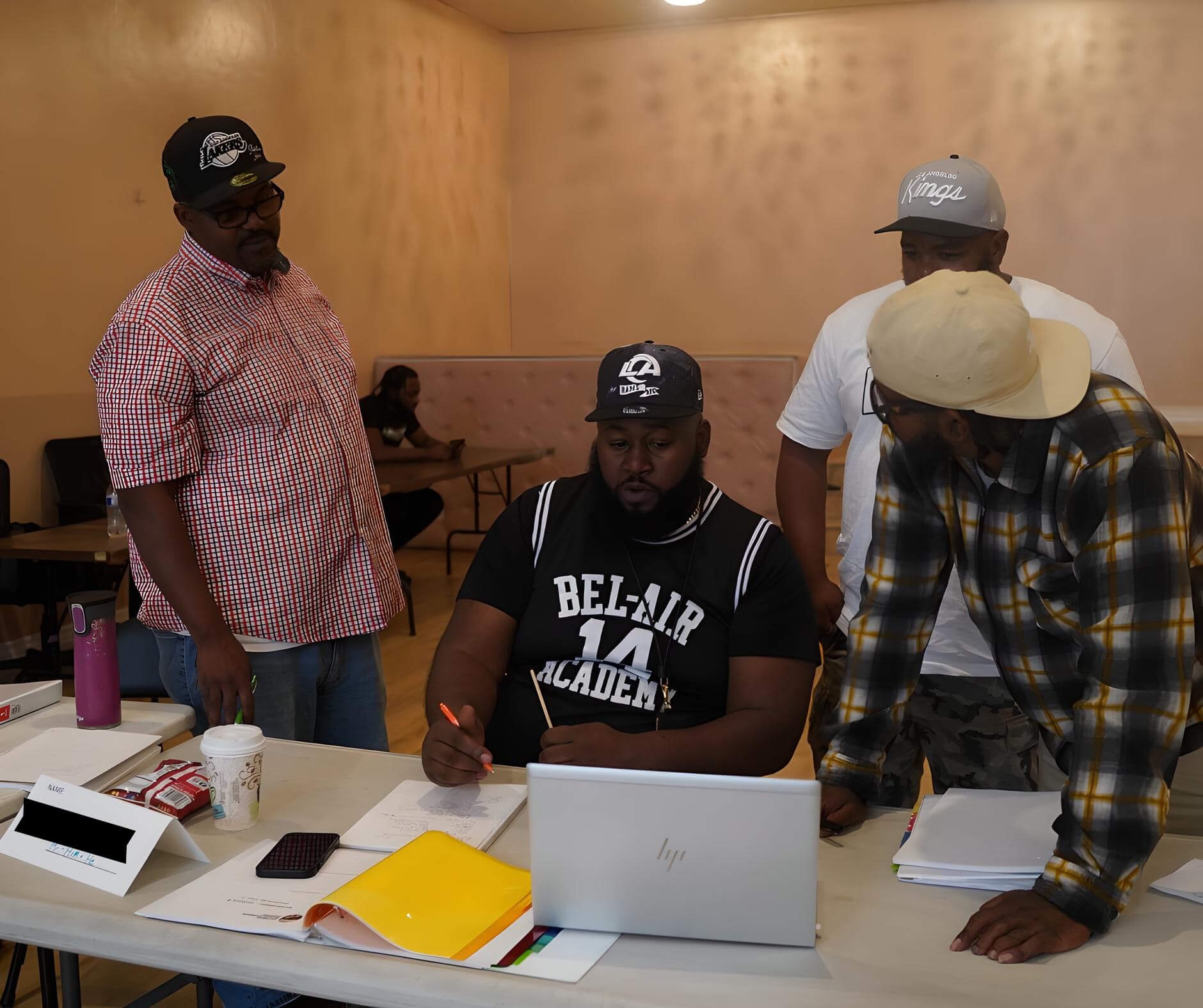 Four men gathered around a table, one seated and three standing. They are engaged in a discussion or activity involving paperwork and a laptop in an indoor setting with wooden-paneled walls. The seated man is writing or drawing, while the others observe.