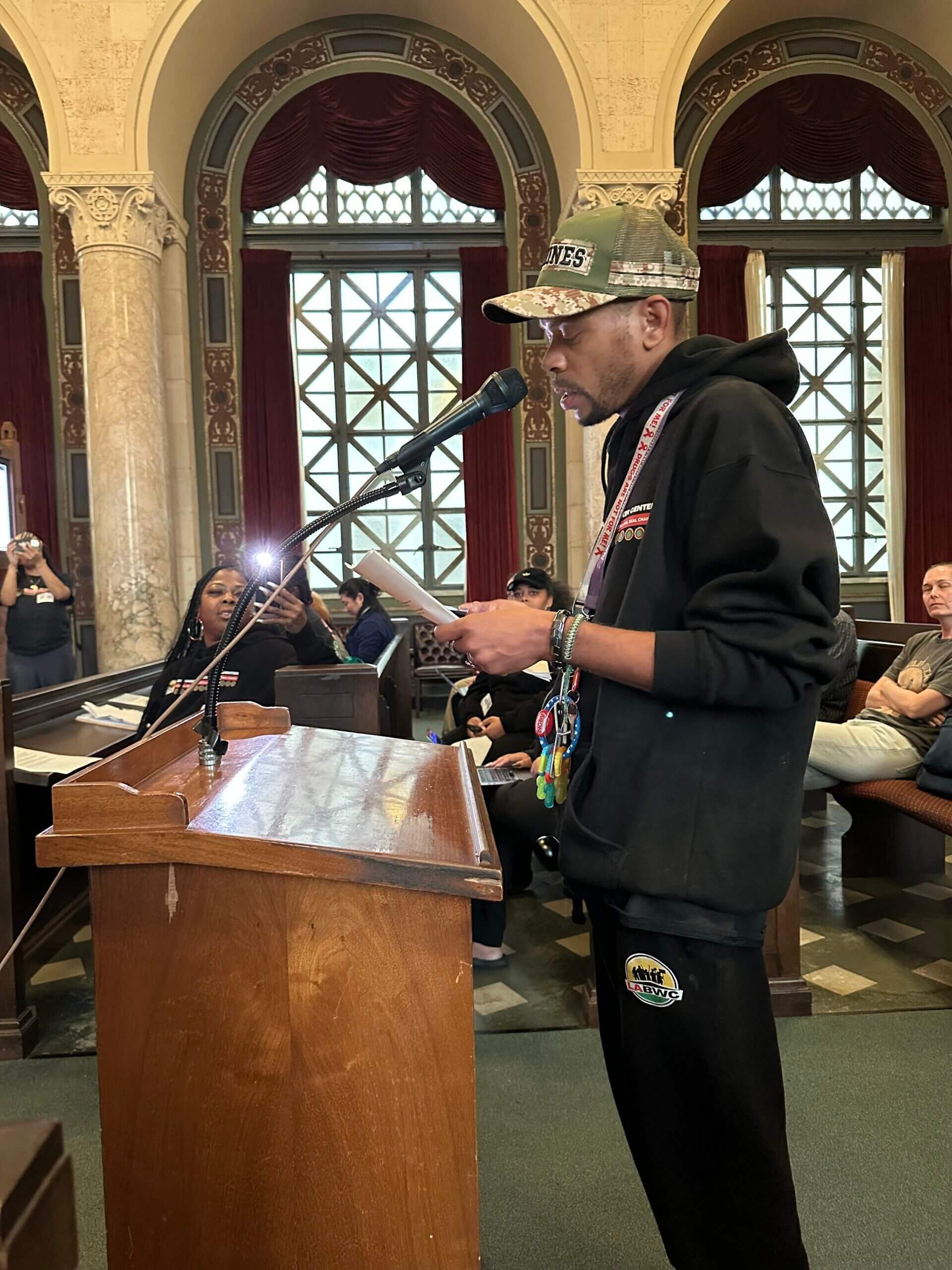 Man in black hoodie and black pants standing at a wooden podium, speaking into a microphone, in a grand hall with high arched windows and ornate interior decoration.