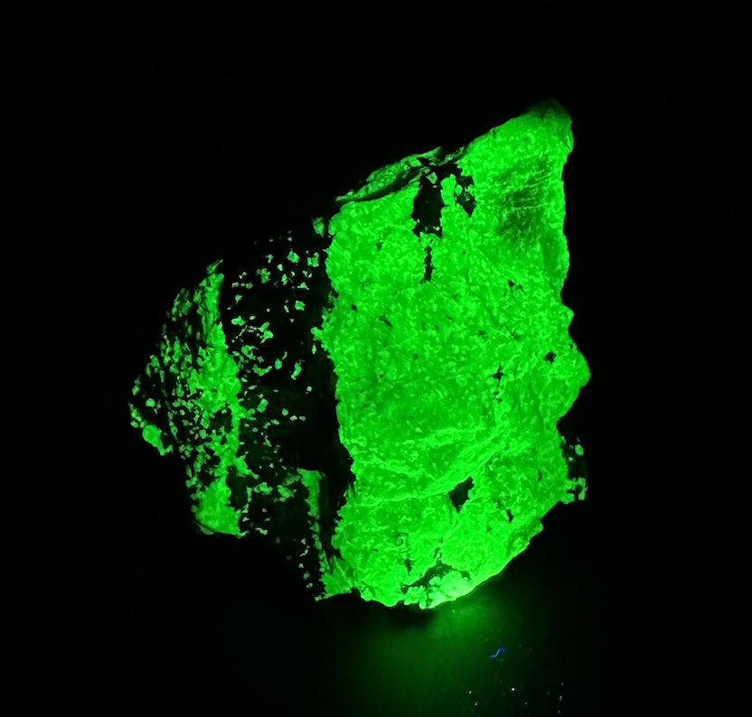 A glowing green mineral or rock emitting light in darkness, with rough, uneven surface and dark patches.