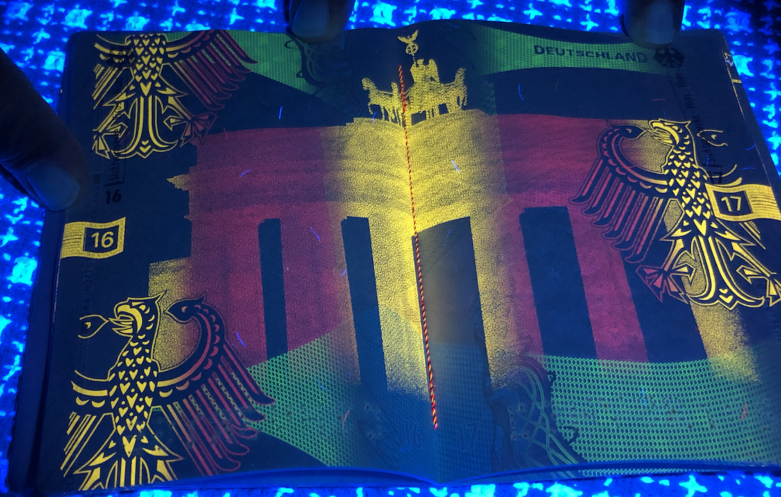 A German passport open to the first page, with the German eagle emblem seen twice, at the top and bottom corners, and a colorful security pattern in the background, illuminated under UV light.
