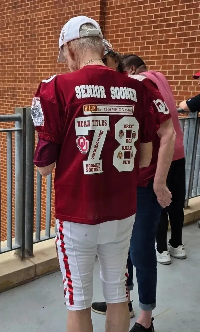 Person wearing a red football jersey with 2022 on the back, standing outdoors next to a metal railing, with other people in the background.