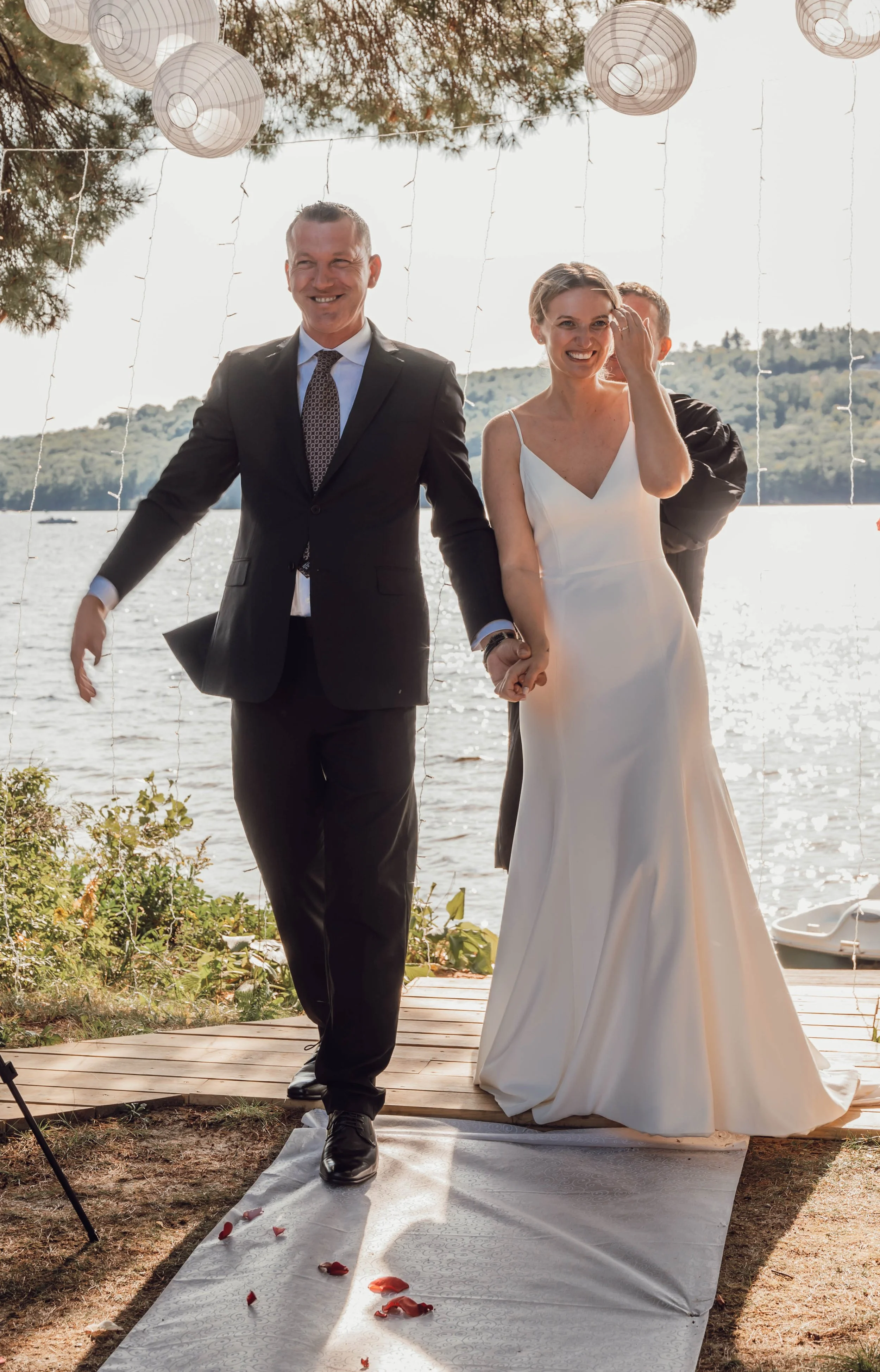 A couple holding hands walking on a white aisle towards a lakeside outdoor wedding altar, with paper lanterns hanging overhead and rose petals scattered on the aisle.
