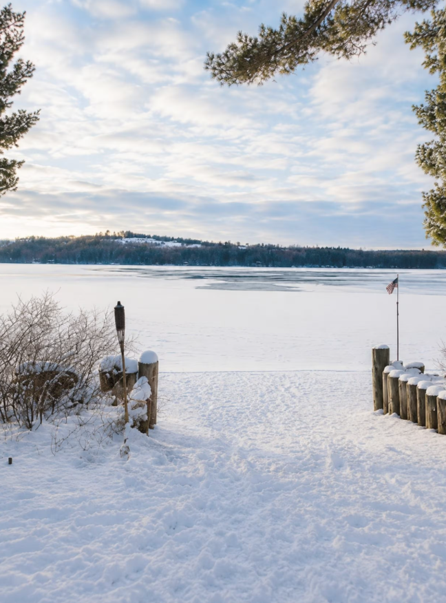Snow-covered pathway leading to a frozen lake, with an American flag on a pole and a small torch on the left, framed by trees with snow on their branches.