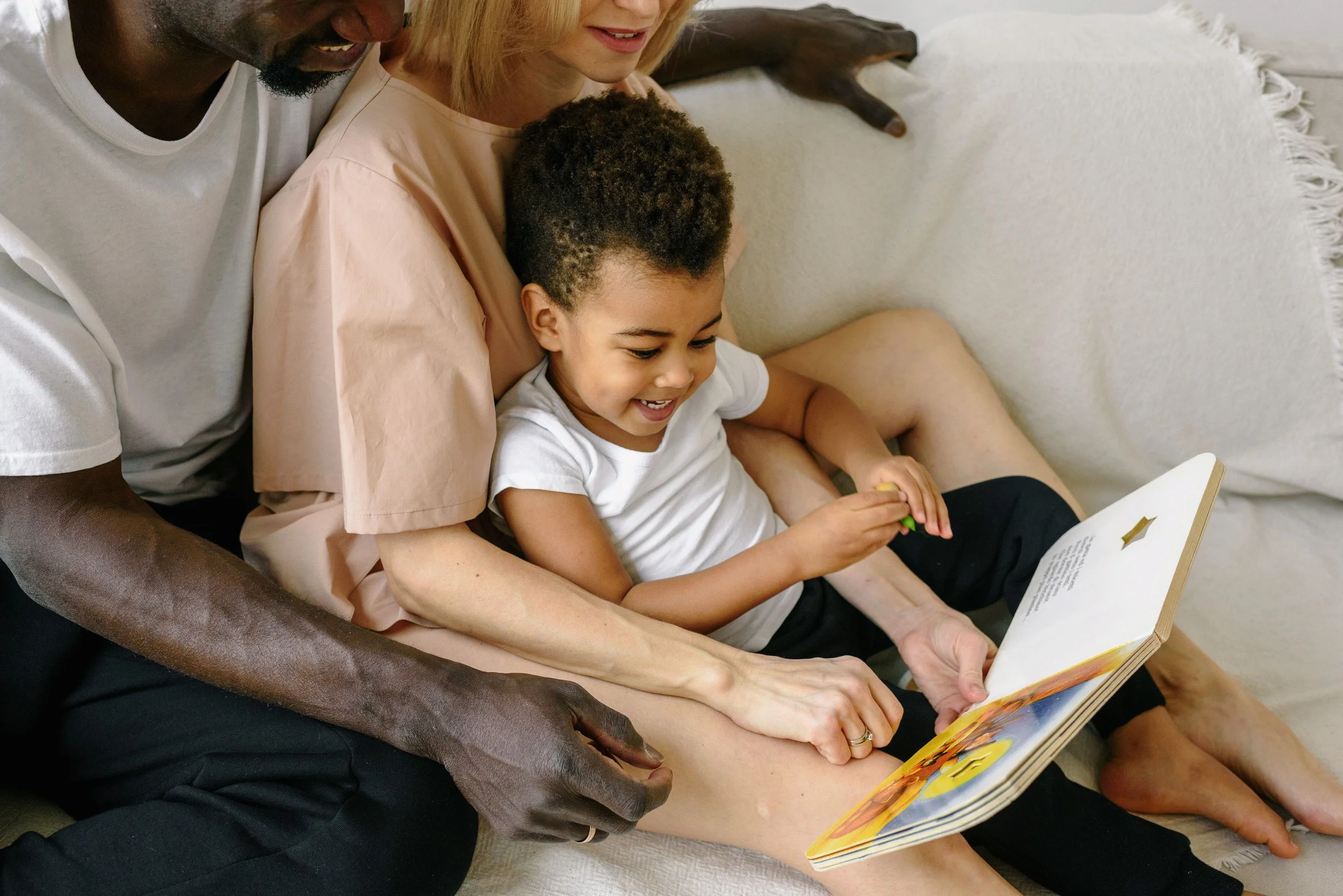A close-up of a family sitting on a white couch, reading a children's book together. The image captures the joy of sharing a story.