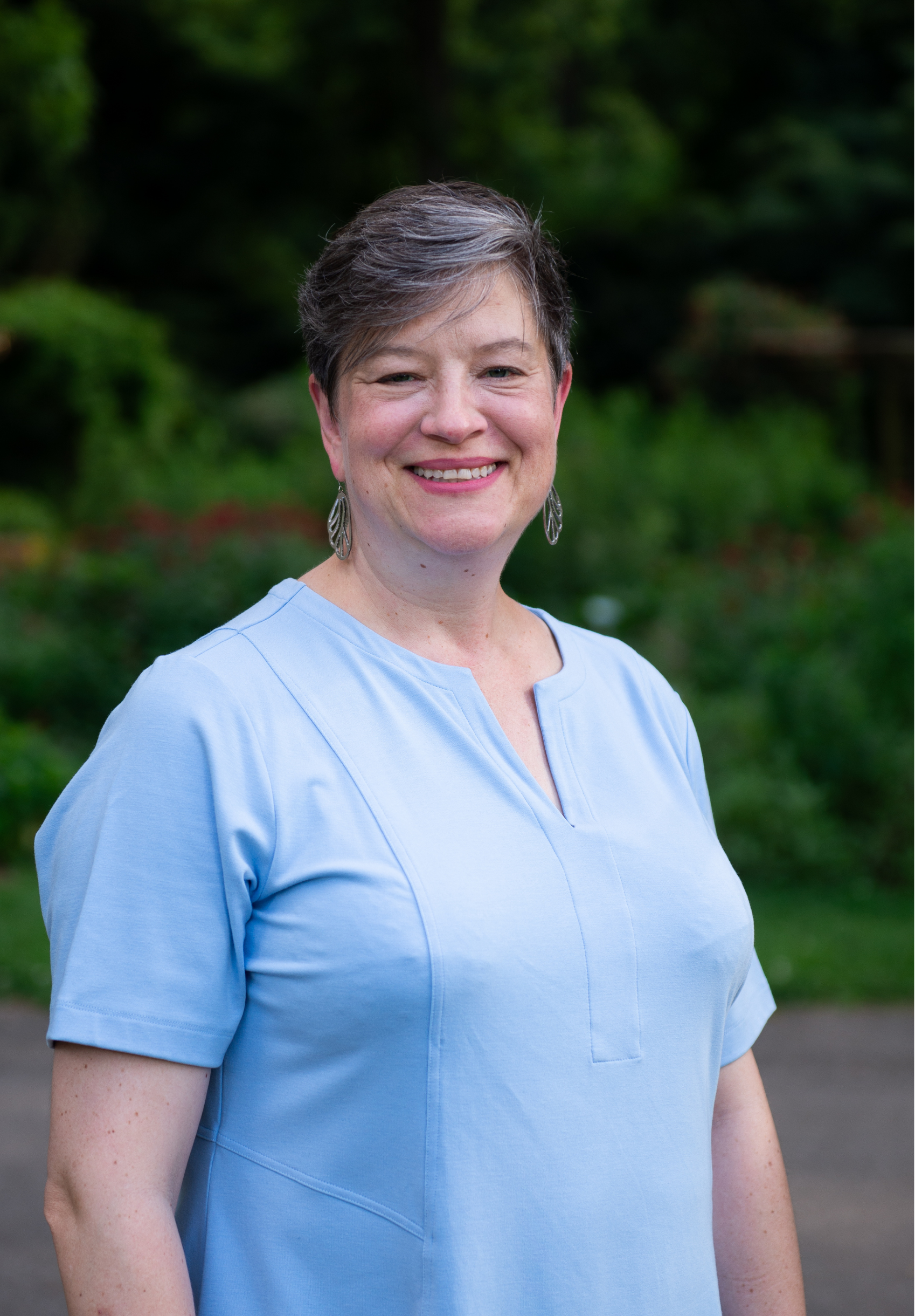 Lisa Gillispie, therapist in Columbus, OH, smiling with short gray hair wearing a light blue top and dangling earrings, standing outdoors with green foliage in the background.