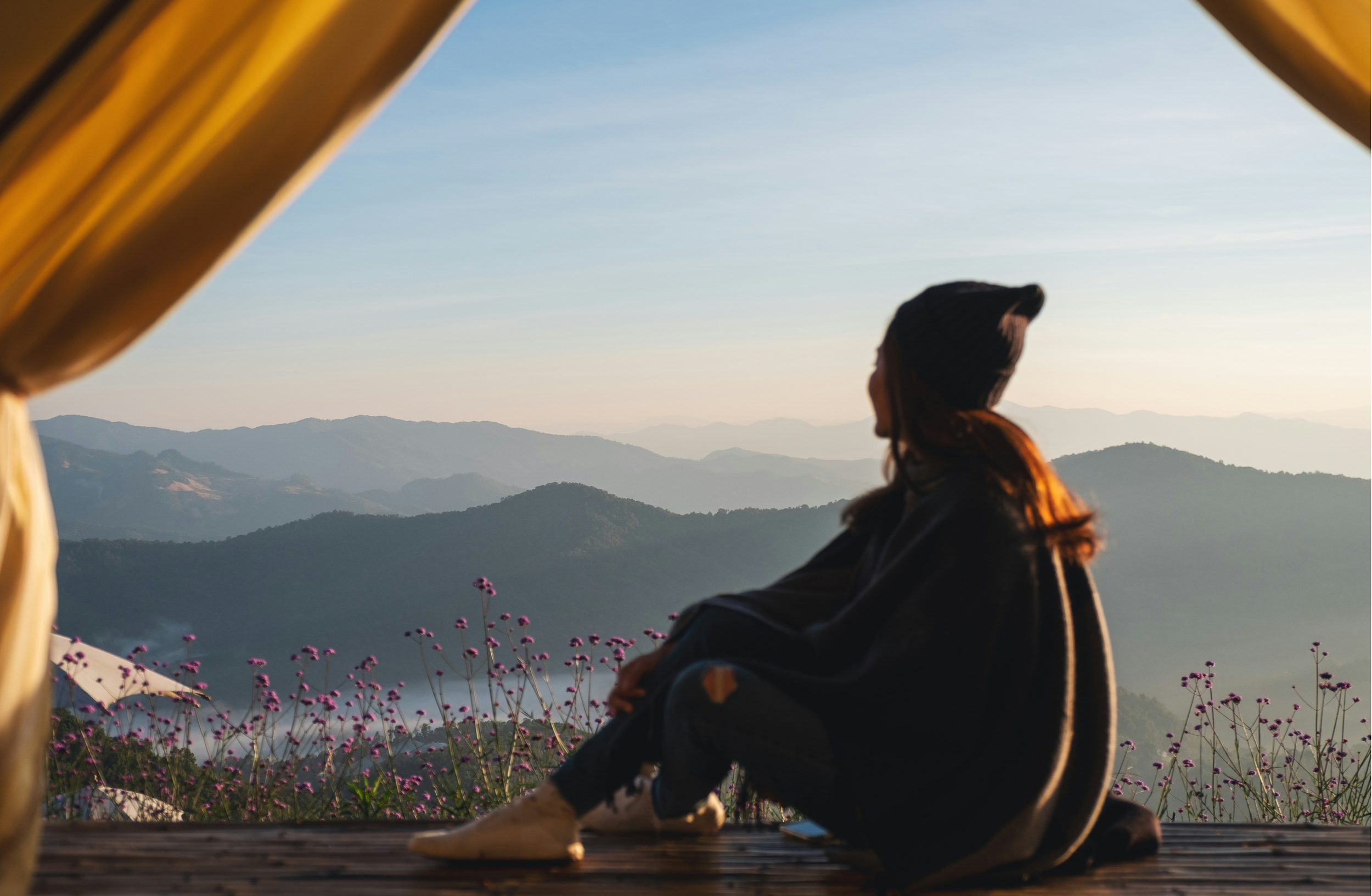 A woman squatting on a wooden platform outdoors, overlooking a mountain range during sunset, with purple flowers in the foreground and a yellow fabric structure framing the scene.