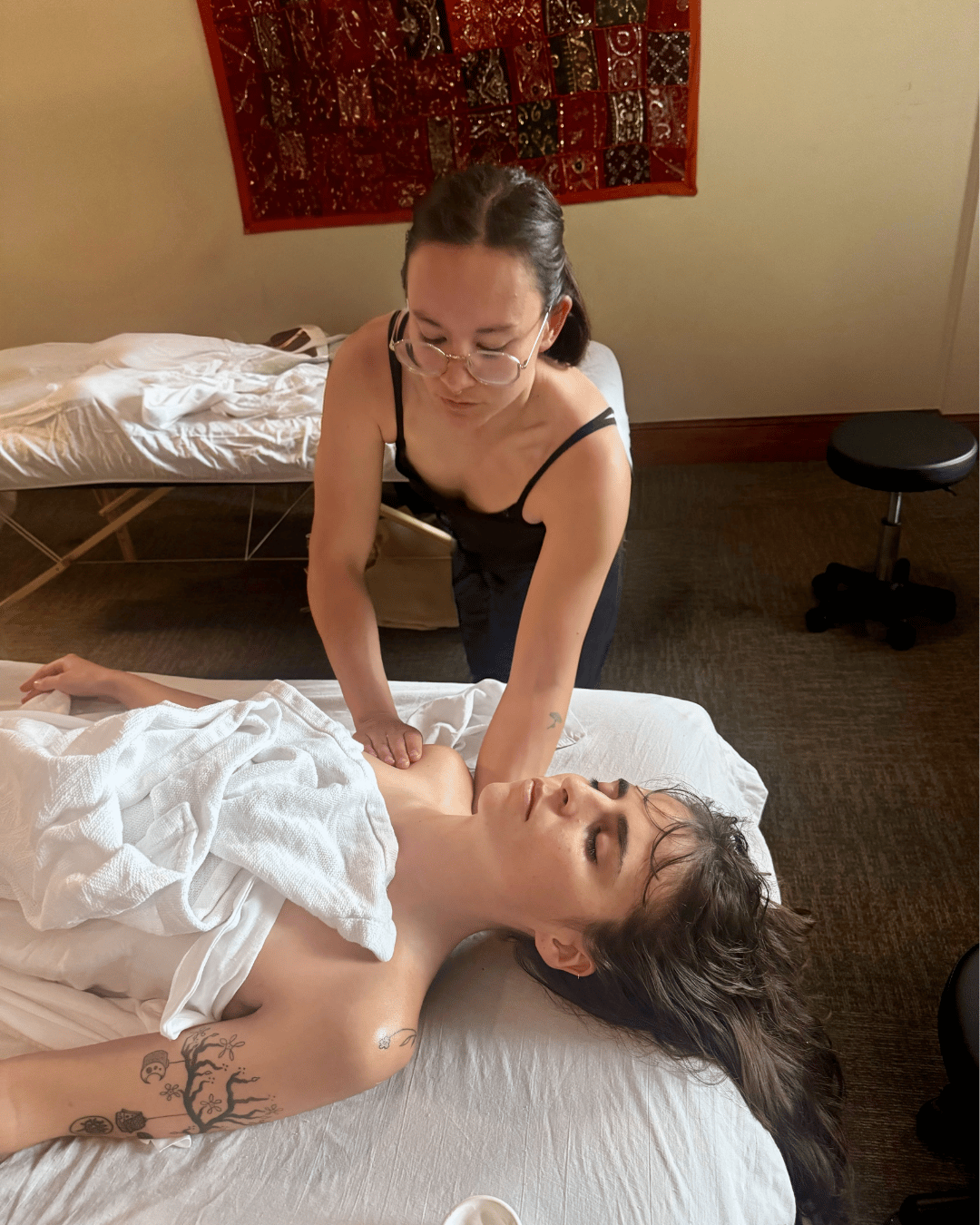 A woman receiving a massage from a therapist in a spa room, with a massage table, white towels, and a decorative textile on the wall.