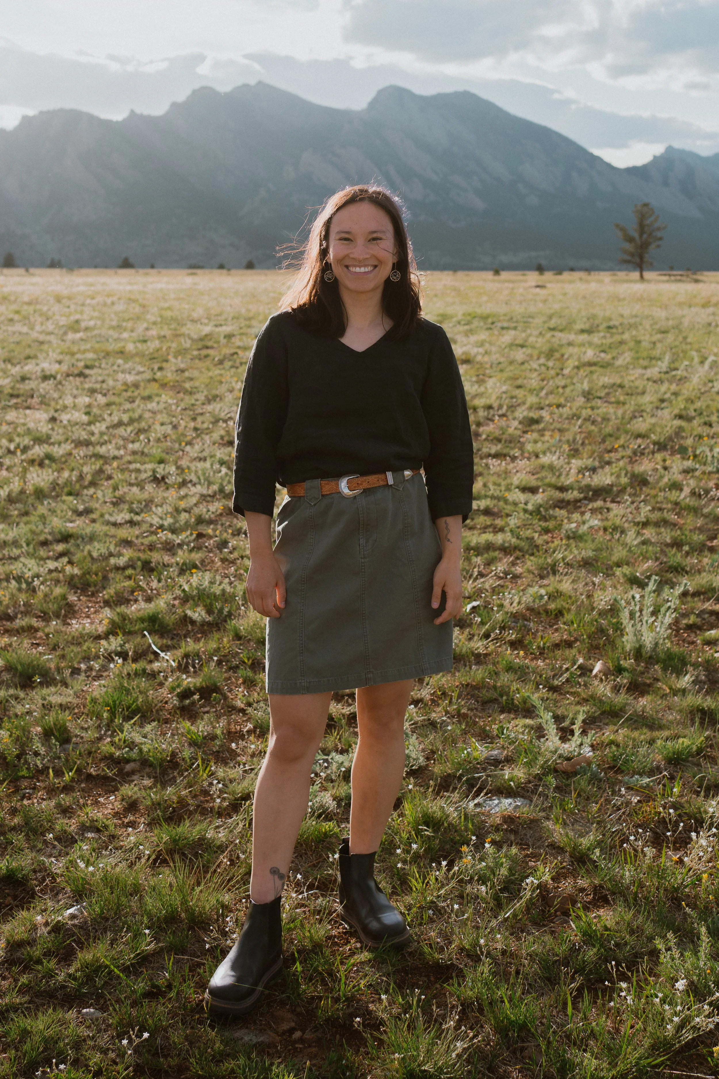 A young woman standing in a grassy field with mountains in the background, smiling at the camera.