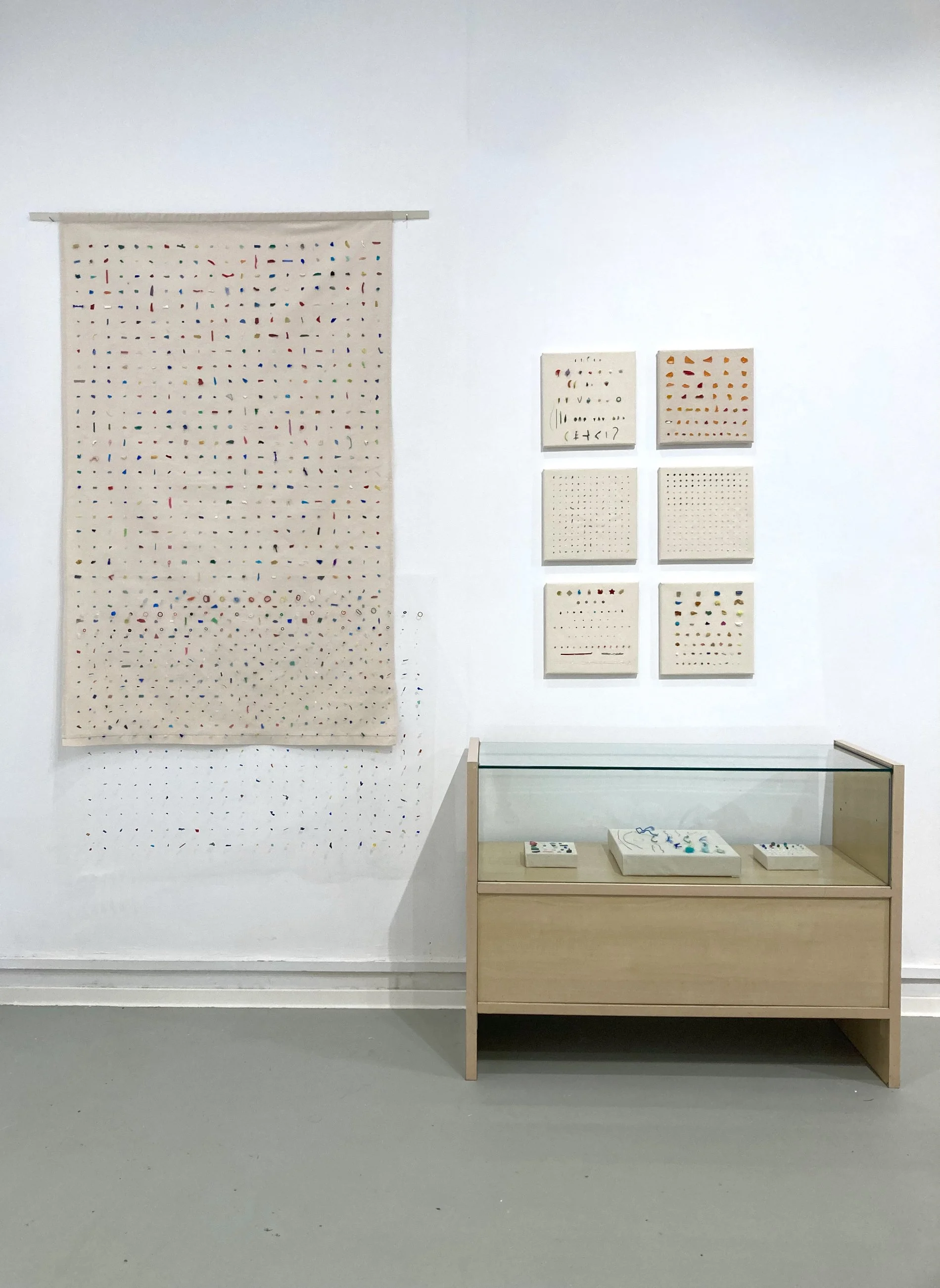 Exposición de arte con obras minimalistas y abstractas en una galería blanca, incluyendo un gran tapiz colgado a la izquierda con patrones coloridos y varias piezas en las paredes, además de una vitrina con muestras en la parte inferior.