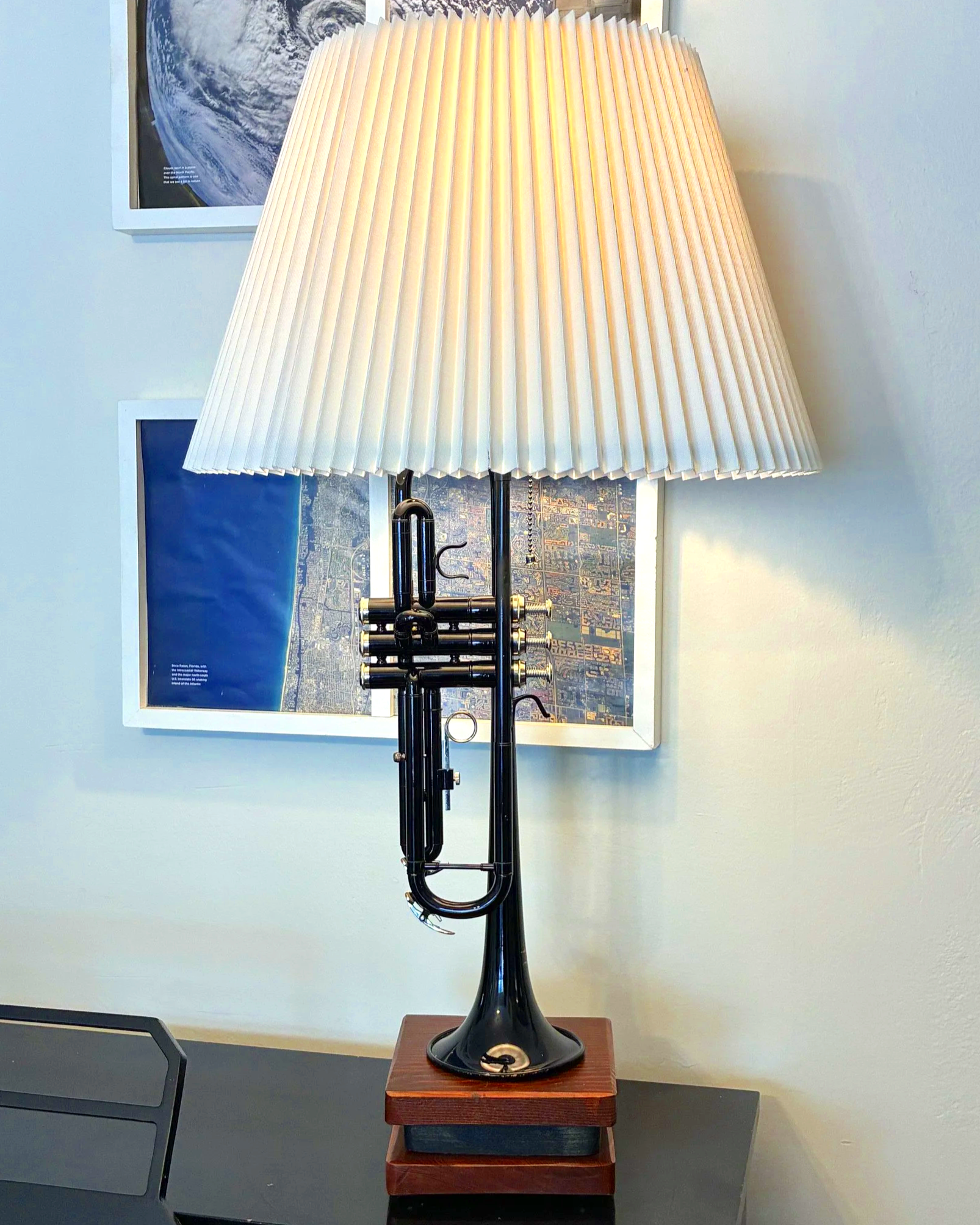 A black trumpet is mounted at the top of a lamp with a large, pleated, white lampshade. The lamp is on a small wooden table against a wall decorated with framed pictures, including a map and a satellite image.