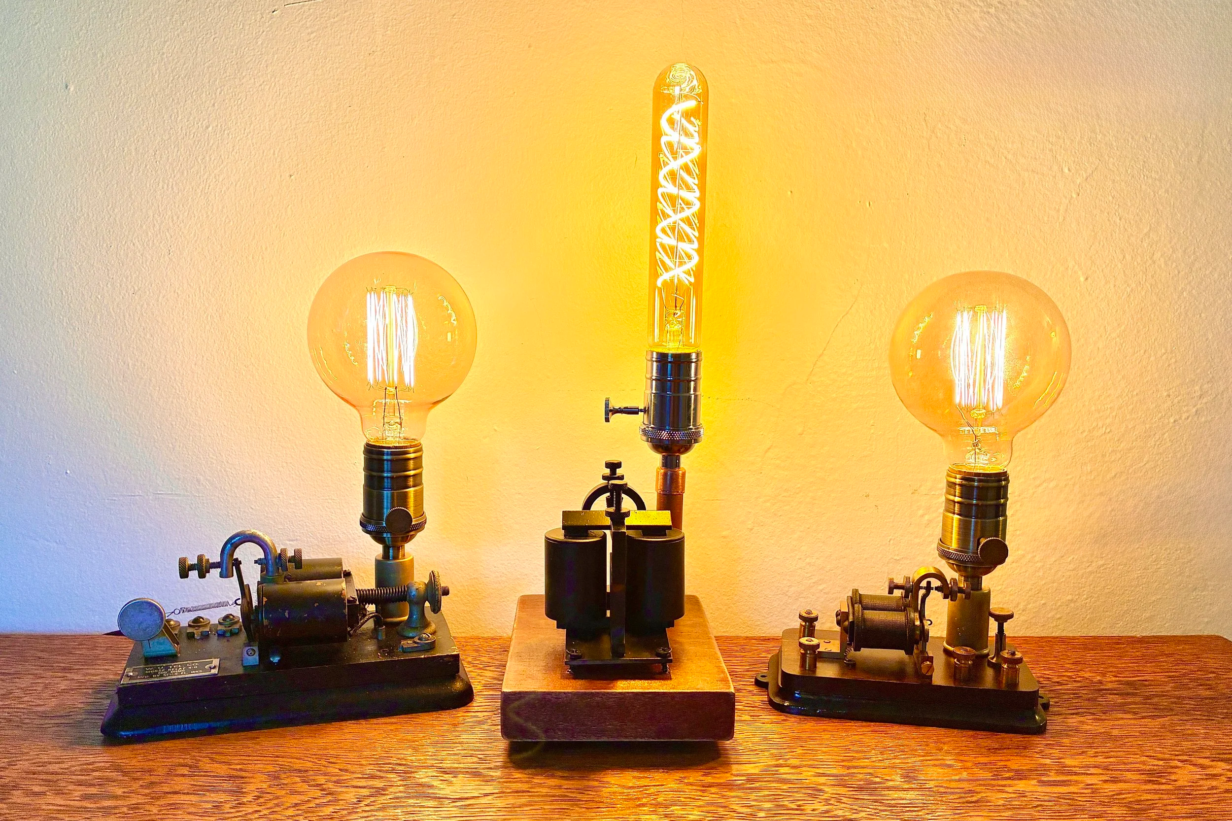 A display of vintage scientific lighting experiment equipment on a wooden surface, featuring three illuminated light bulbs with filament designs, mounted on metallic bases with adjustable knobs and connectors.