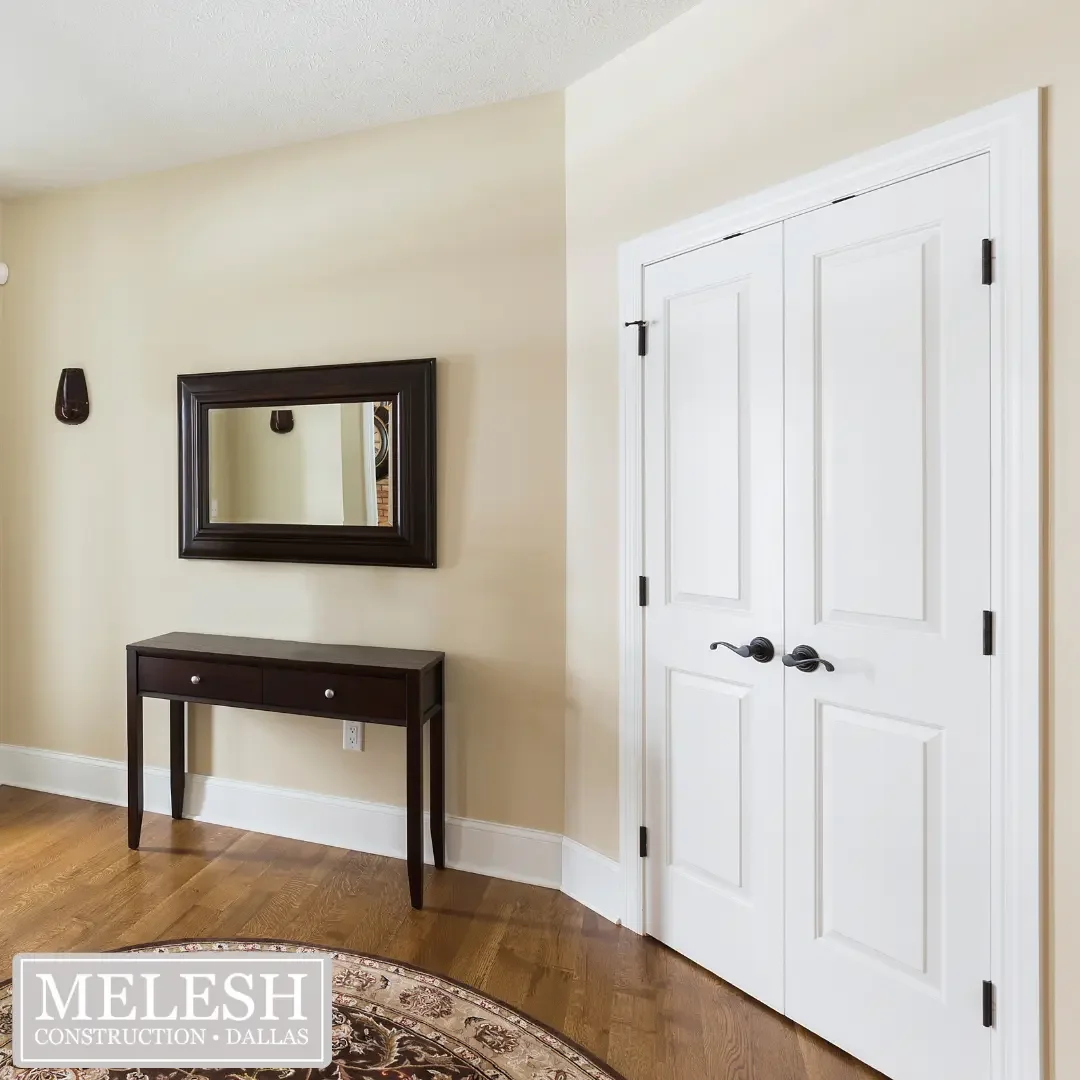 Interior of a room with double white doors, a black framed mirror, a small black console table, a wood floor, and cream-colored walls.