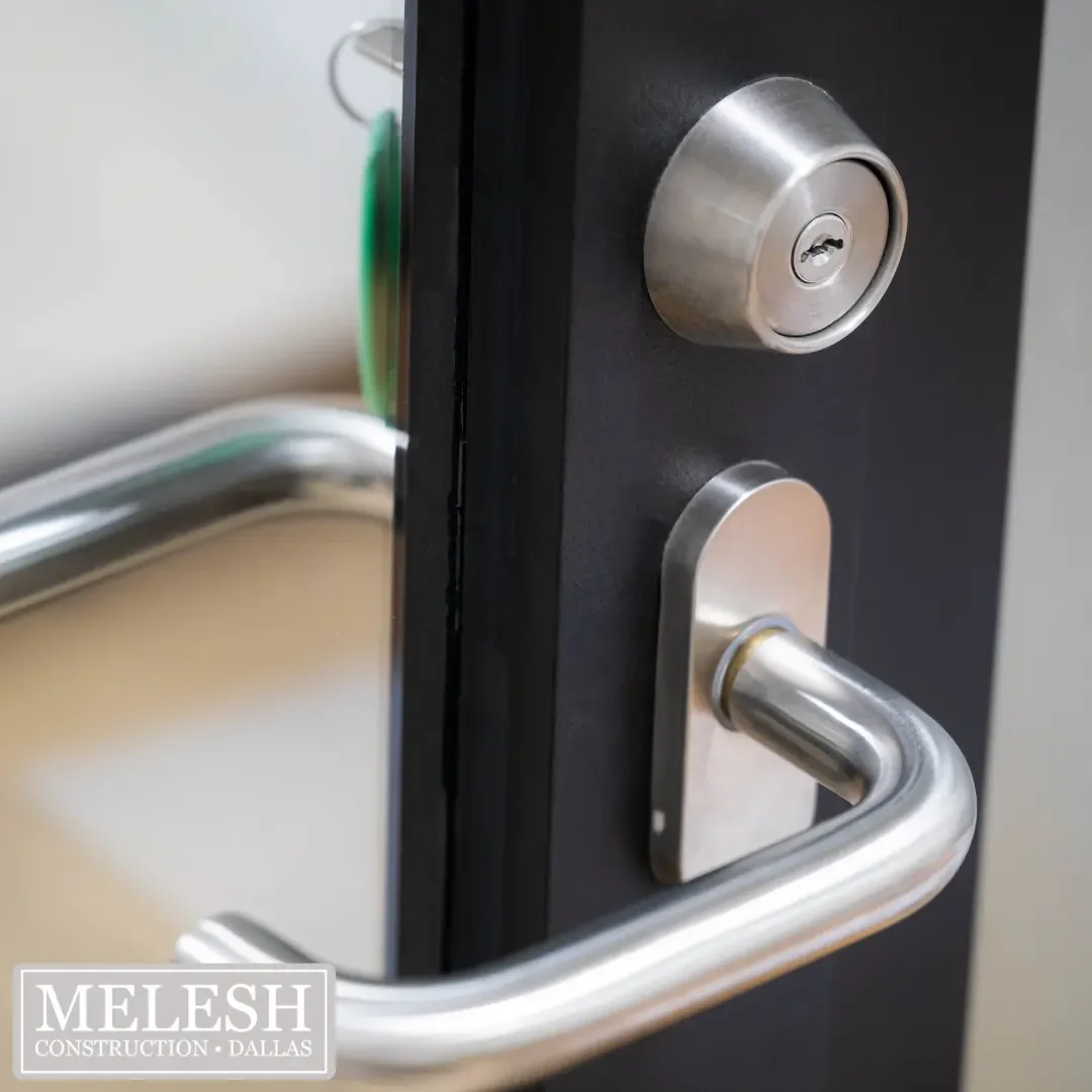 Extreme close-up of a brushed stainless steel keyed deadbolt and lever door handle mounted on a dark navy door, representing the accessible lever hardware that Melesh Construction Dallas specifies for wheelchair-accessible doorways