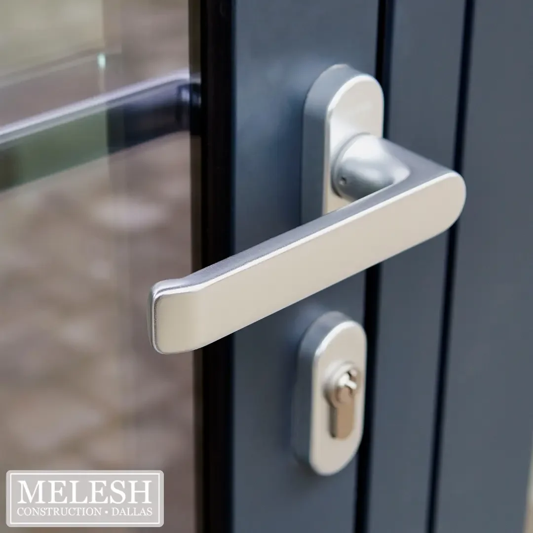Close-up of a brushed nickel flat-bar lever door handle with a rounded keyed lock cylinder mounted on a dark charcoal door with a narrow glass sidelight panel, representing the accessible lever hardware specification