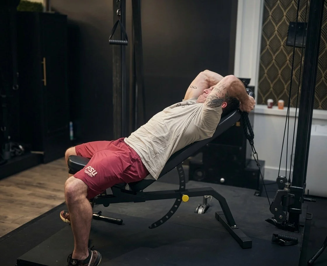 A man lying on a workout bench with his hands behind his head, in a gym setting, preparing for an exercise.