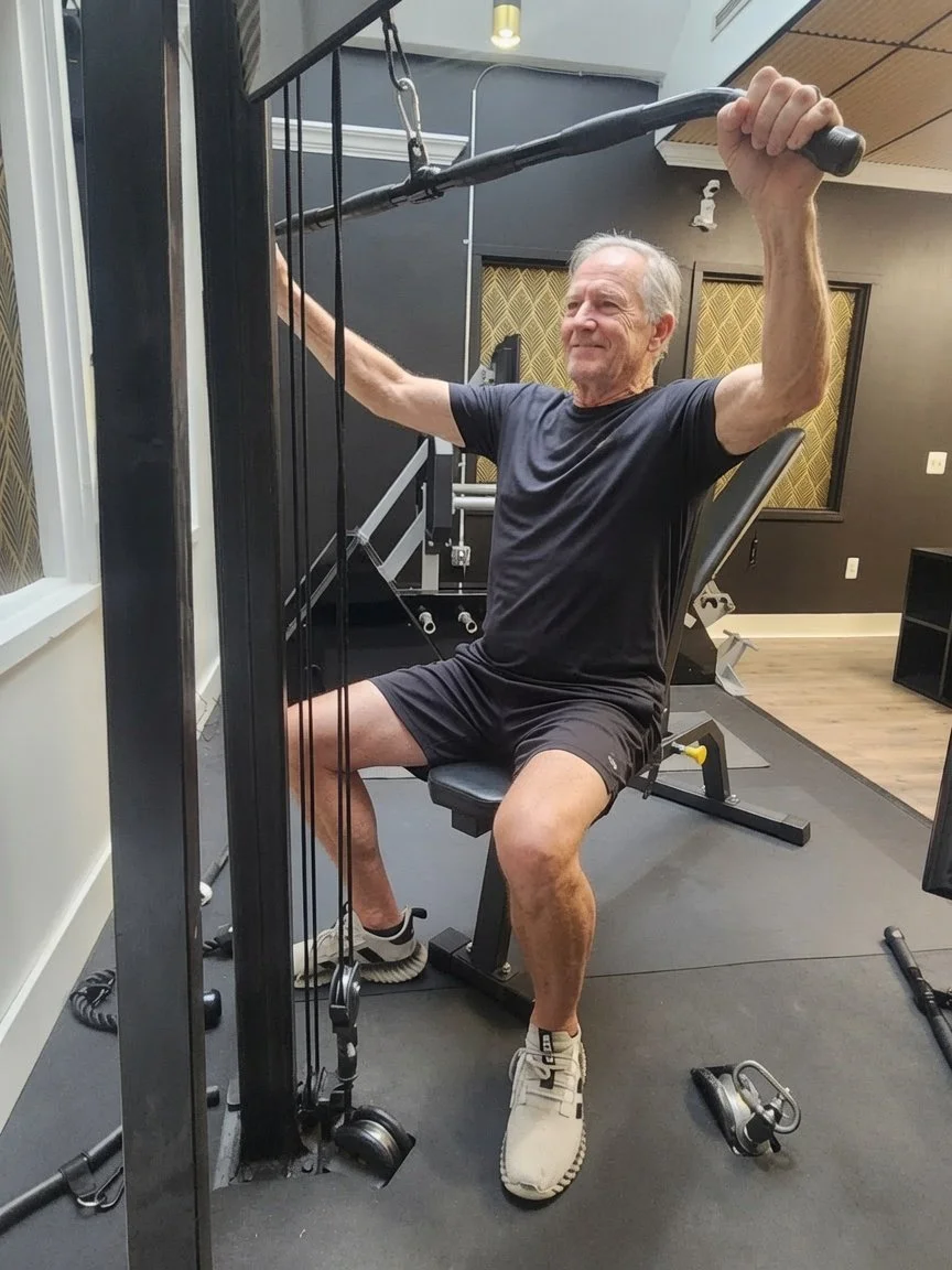 An older man exercising on a gym machine, using a pulley system with a handlebar while sitting on a bench in a gym.