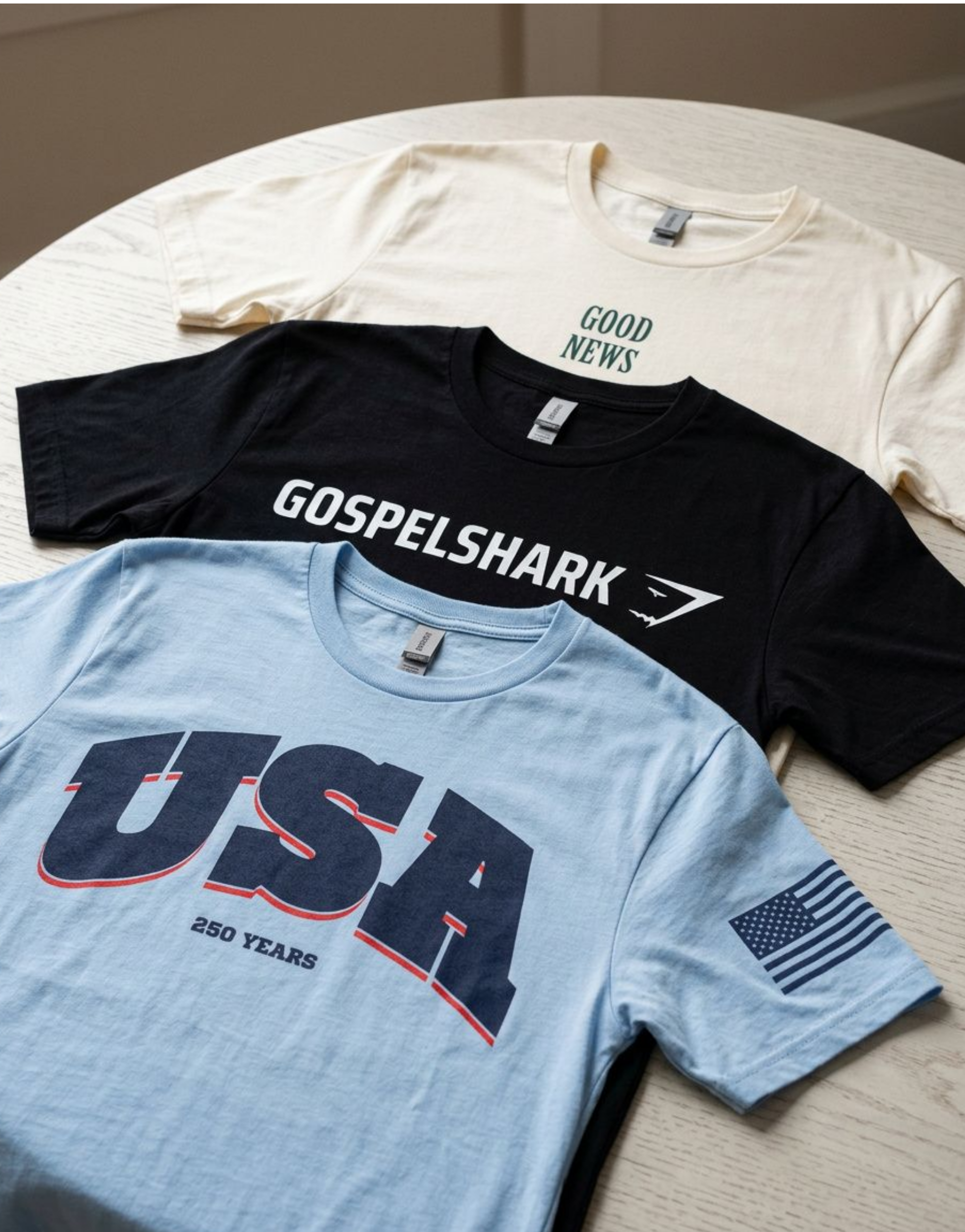Three T-shirts laid flat on a light-colored wooden table, one cream with 'GOOD NEWS' text, one black with 'GOSPEL SHARK' text and logo, and one light blue with a patriotic American flag design and text celebrating 250 years.
