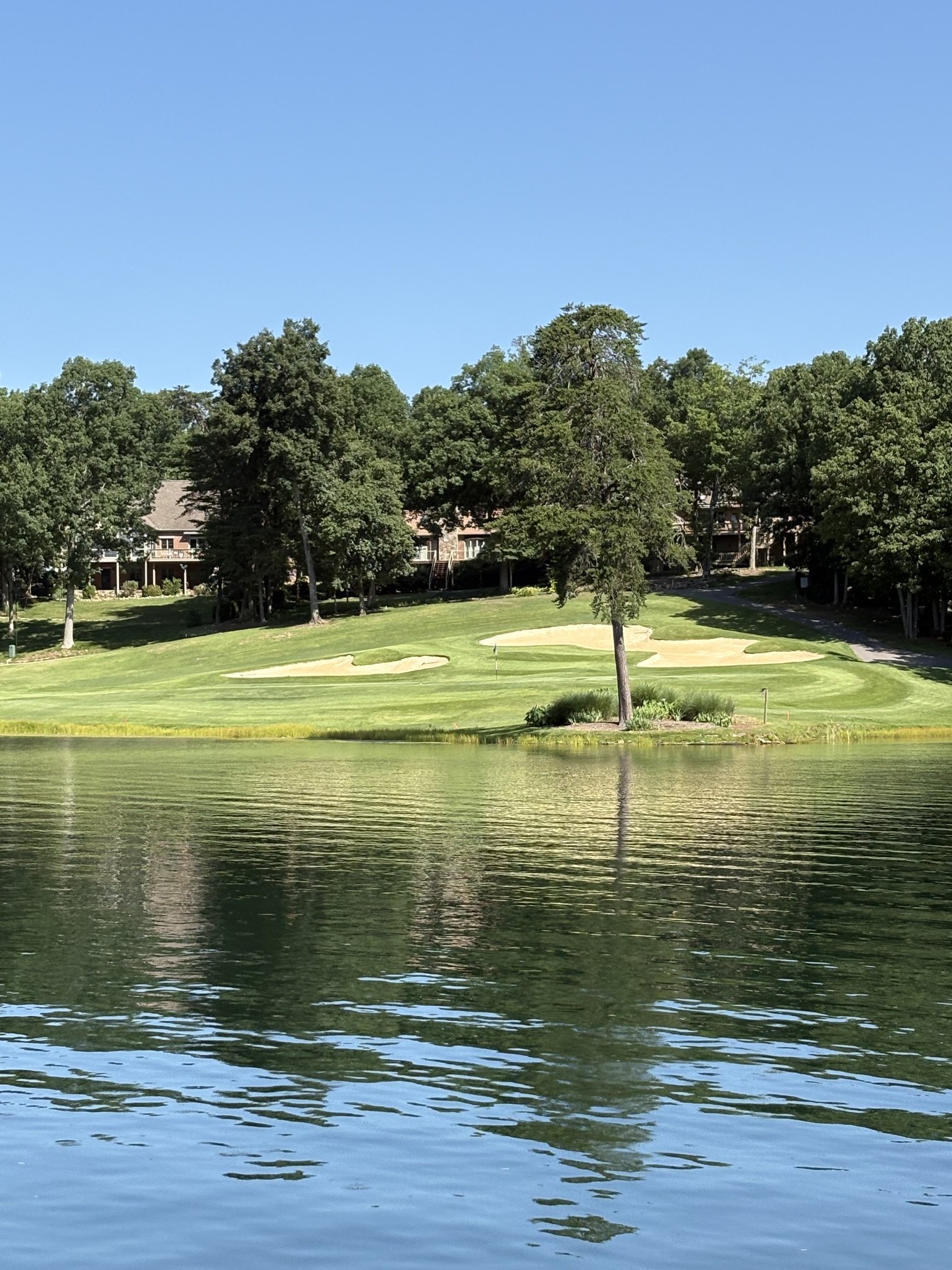 A scenic view of a golf course in Fairfield Glade with a pond in the foreground, grassy areas, and numerous trees with houses visible in the background under a clear blue sky.