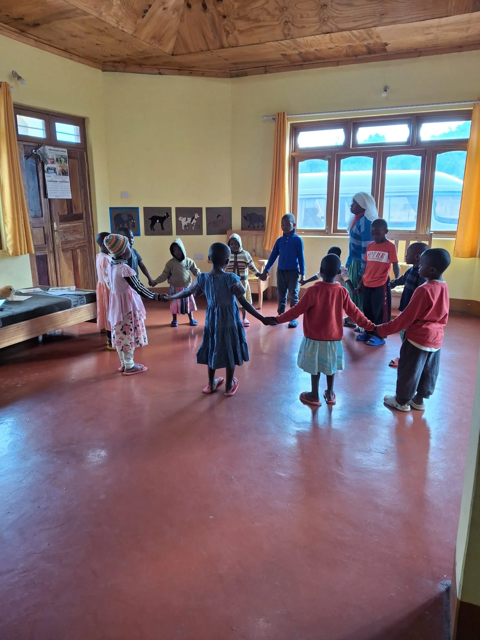 Children hold hands in a circle in a classroom with wooden ceiling, yellow walls, and large windows, decorated with animal posters.