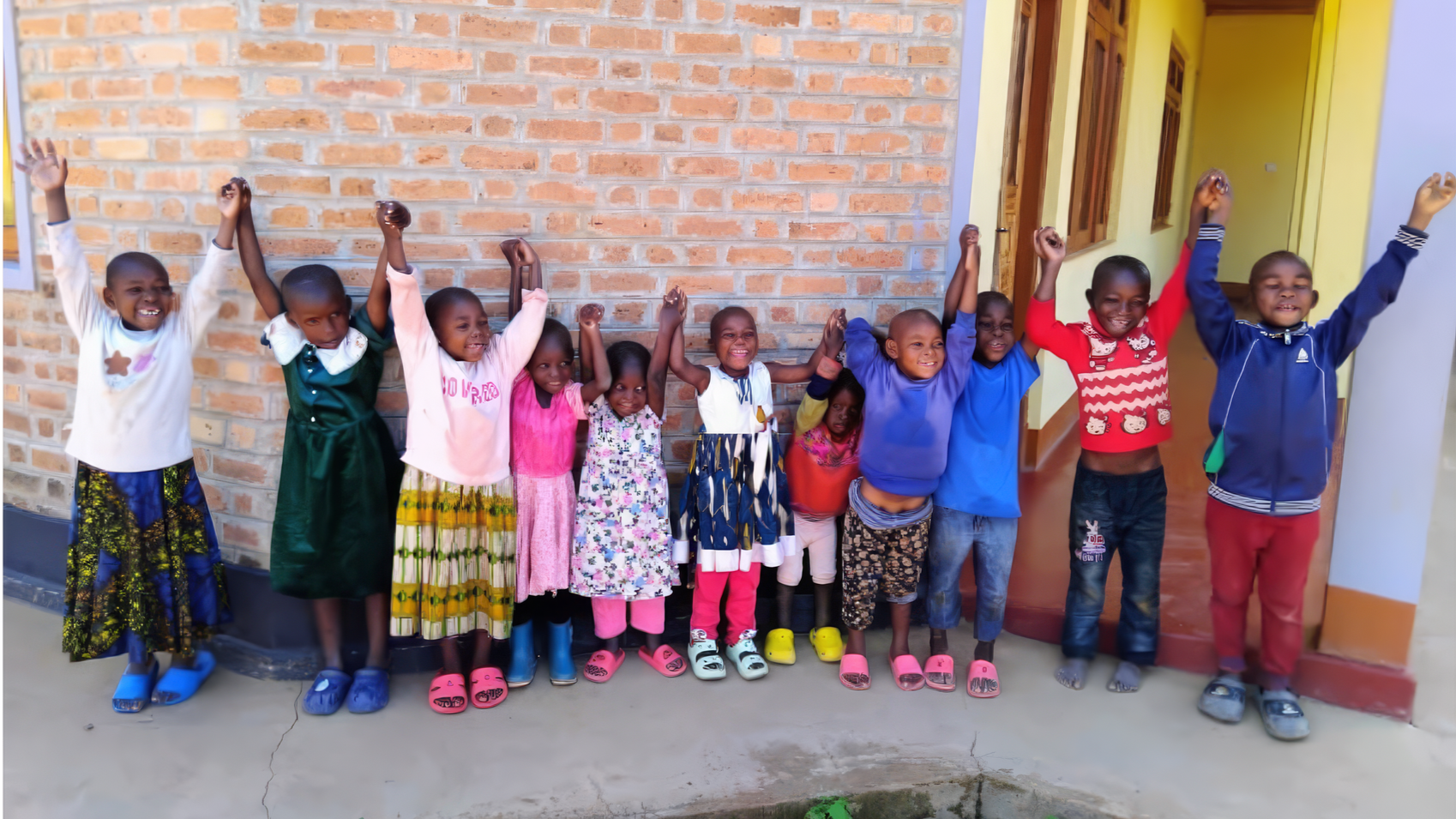 A group of young children standing indoors against a brick wall, holding their hands up in the air, smiling and appearing happy.
