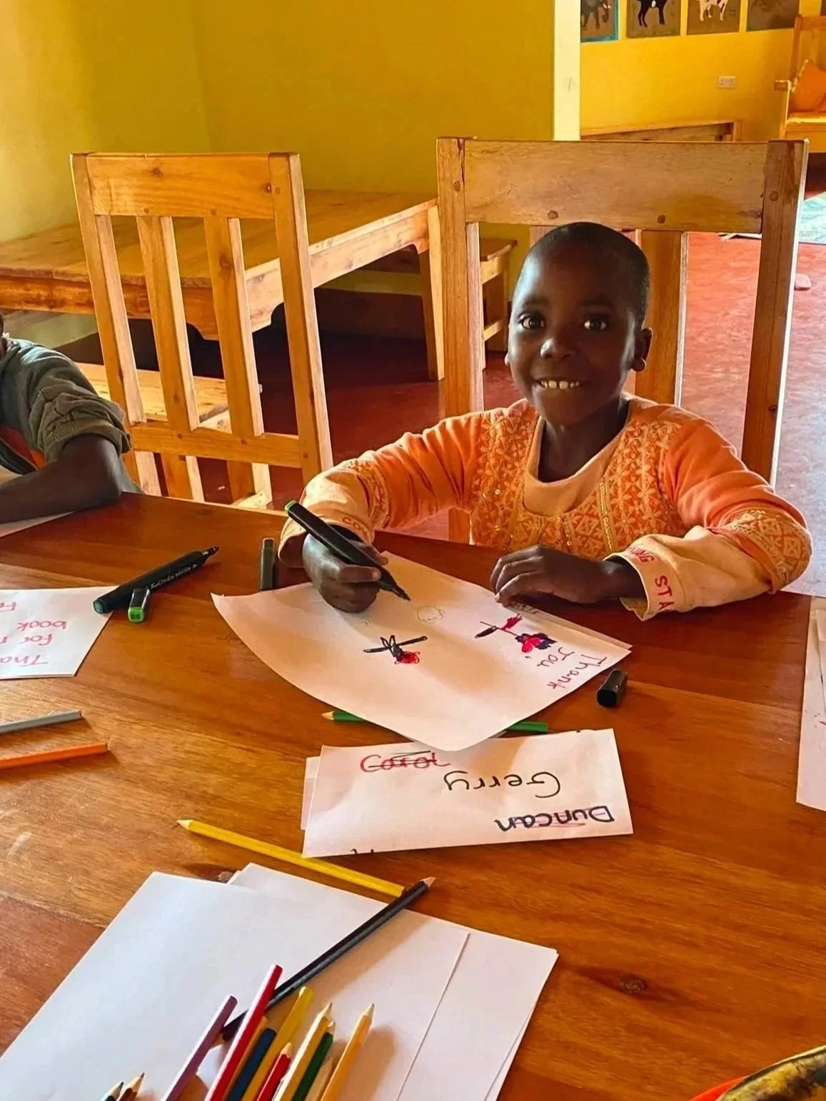 A young girl sitting at a wooden table, smiling and holding a black marker, with drawings and words about traffic safety on her paper, in a classroom with yellow walls and wooden furniture.
