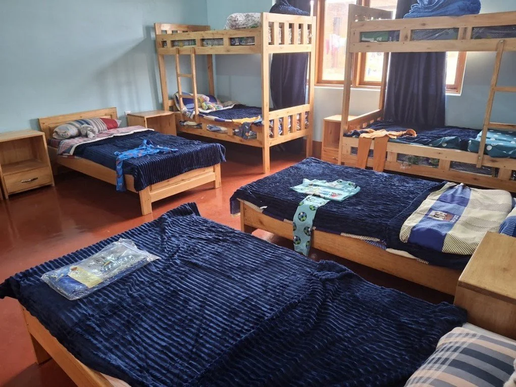A room with five wooden beds, including two bunk beds, all with blue blankets and pillows. There are two wooden nightstands and a window with blue curtains.