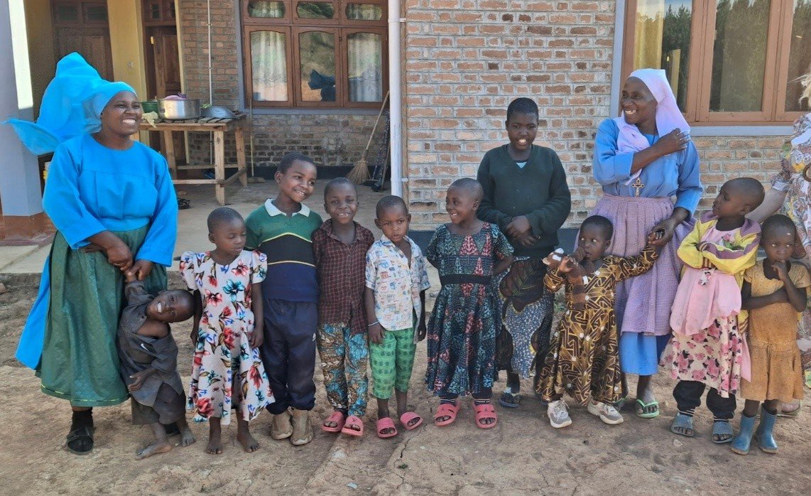 Group of children and two women standing outside a brick building, some of the children are smiling and wearing colorful clothes, one woman in blue is smiling and wearing a large blue headwrap, the other woman is dressed in blue and a pink headscarf.