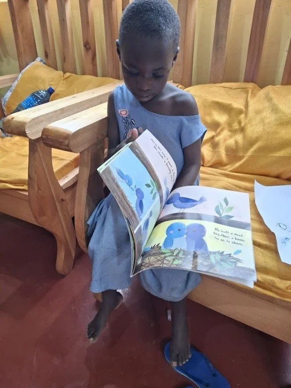 A young child sitting on a bed, reading a colorful children's book about bluebirds, with a wooden chair beside the bed and a water bottle nearby.