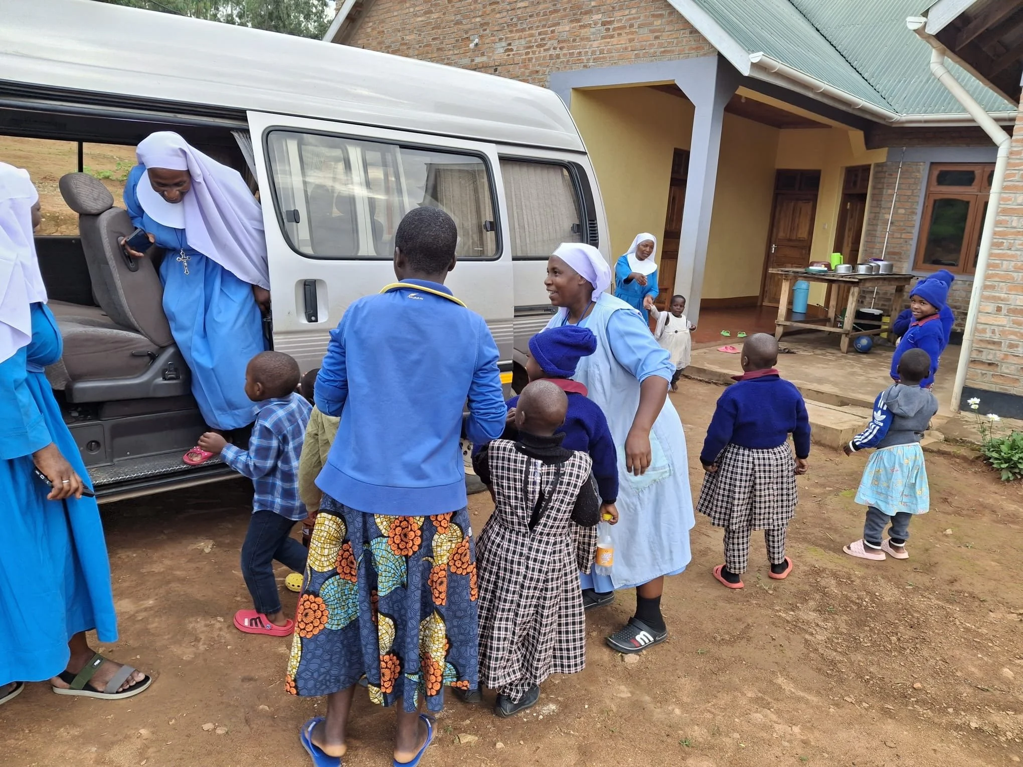 Group of children and women with light to dark brown skin gathering outside near a house with a brick exterior and metal roof, around a white van, on a dirt ground.
