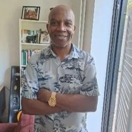 A smiling man with a bald head, wearing a patterned short-sleeve shirt and a gold wristwatch, standing with arms crossed indoors near a window and bookshelf.